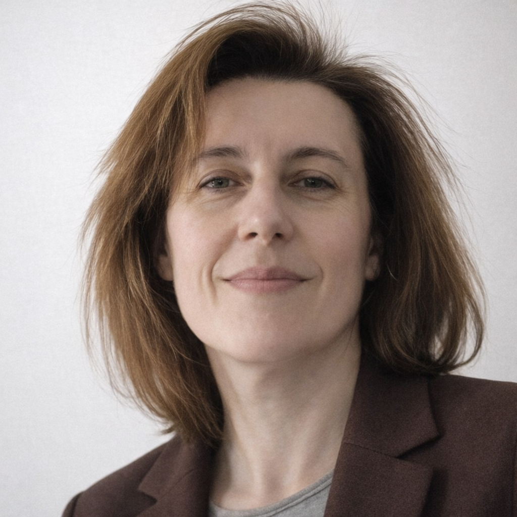 A woman with shoulder-length auburn hair wearing a dark blazer and light-colored top, smiling slightly, against a plain light background.