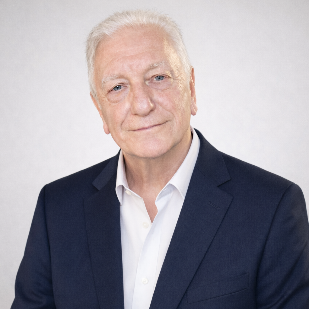 An older man with white hair and blue eyes wearing a navy blazer and white shirt, smiling softly against a plain light background.