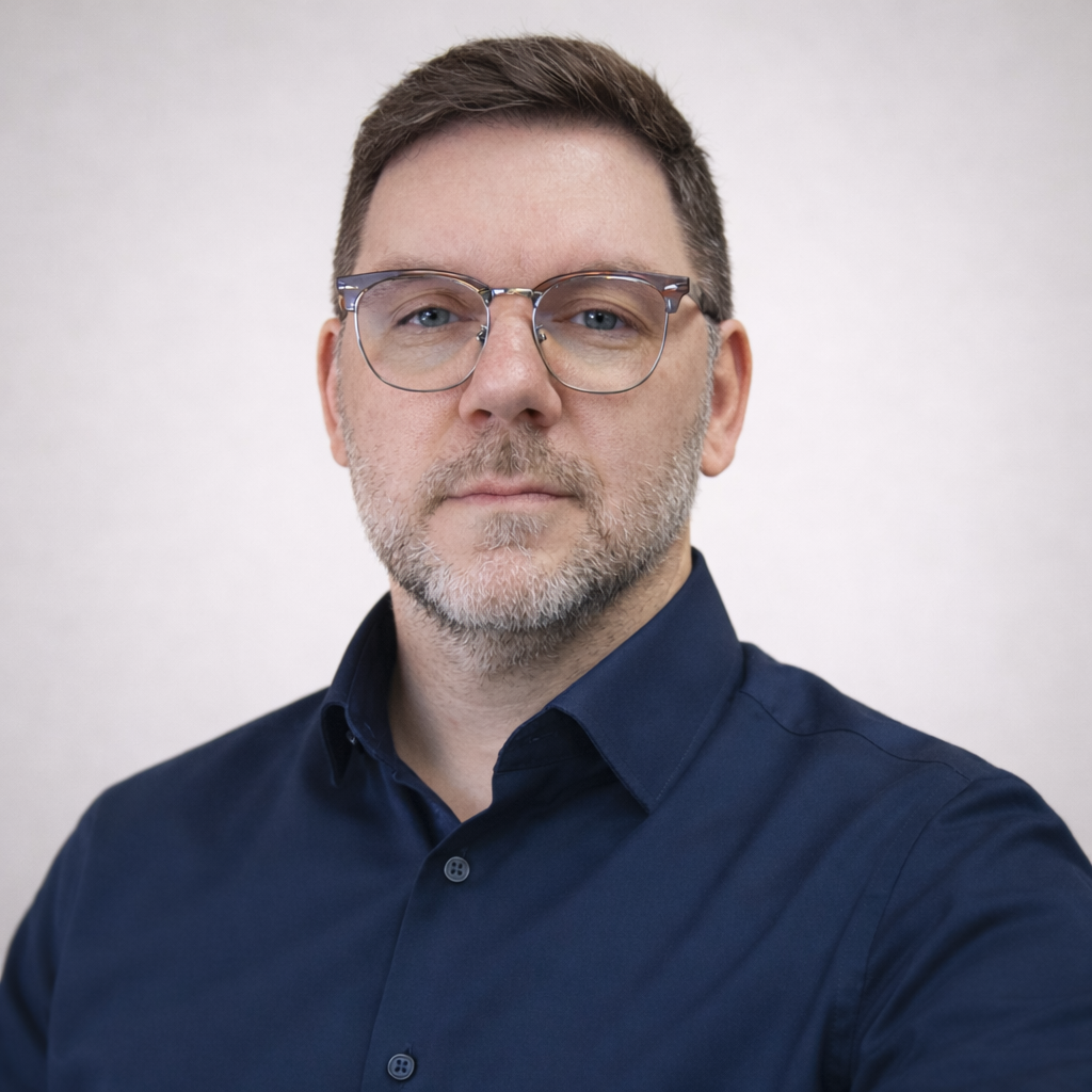 A middle-aged man with short brown hair, glasses, and a beard wearing a dark blue button-down shirt, looking directly at the camera with a neutral expression against a plain light gray background.