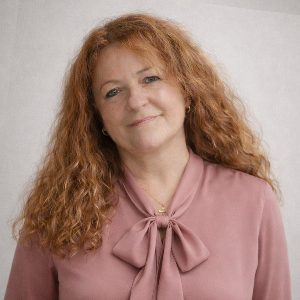 A woman with long curly red hair and light skin wearing a pink blouse with a bow tie collar, standing against a plain background.