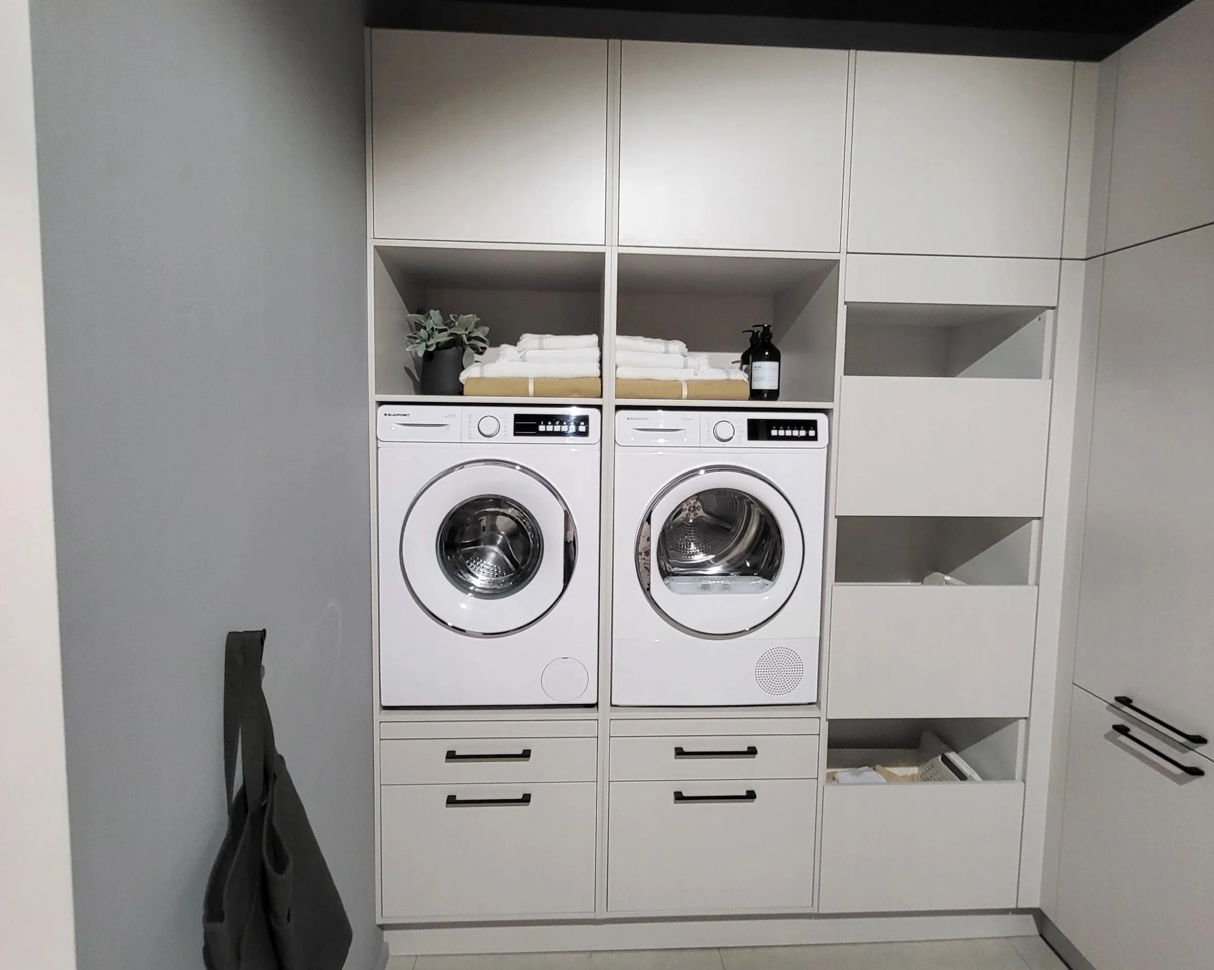 Laundry room with white washing machine and dryer, shelves with folded towels, plants, and cleaning supplies.