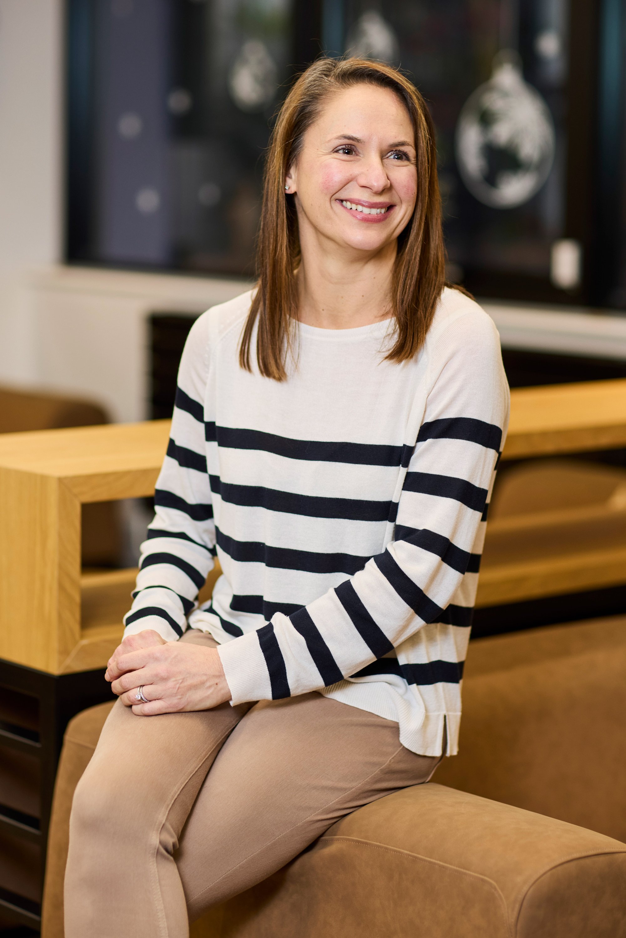A woman with shoulder-length brown hair wearing a white and black striped long-sleeve shirt and beige pants, sitting on a tan leather couch in a modern indoor setting.