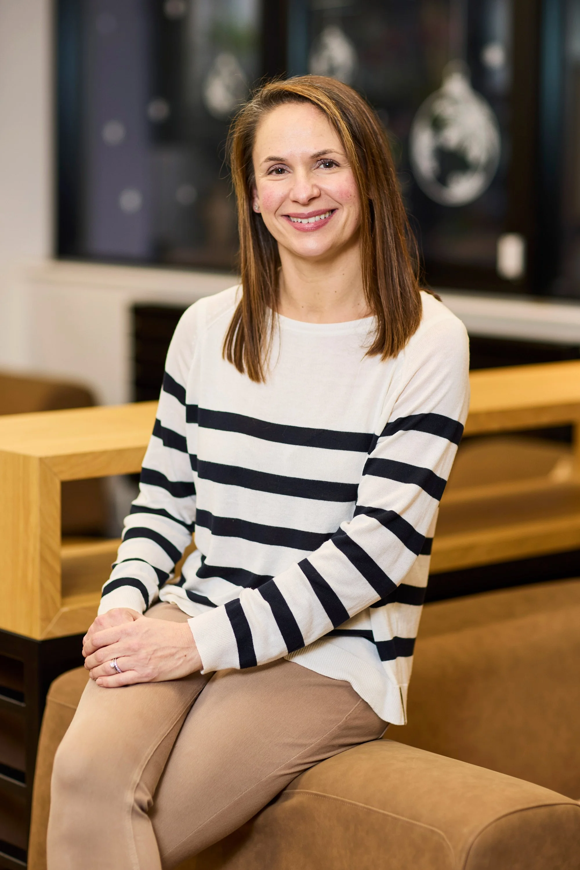 A woman with shoulder-length brown hair smiling and sitting on a brown couch in a modern indoor setting, wearing a white and black striped long-sleeve shirt and beige pants.