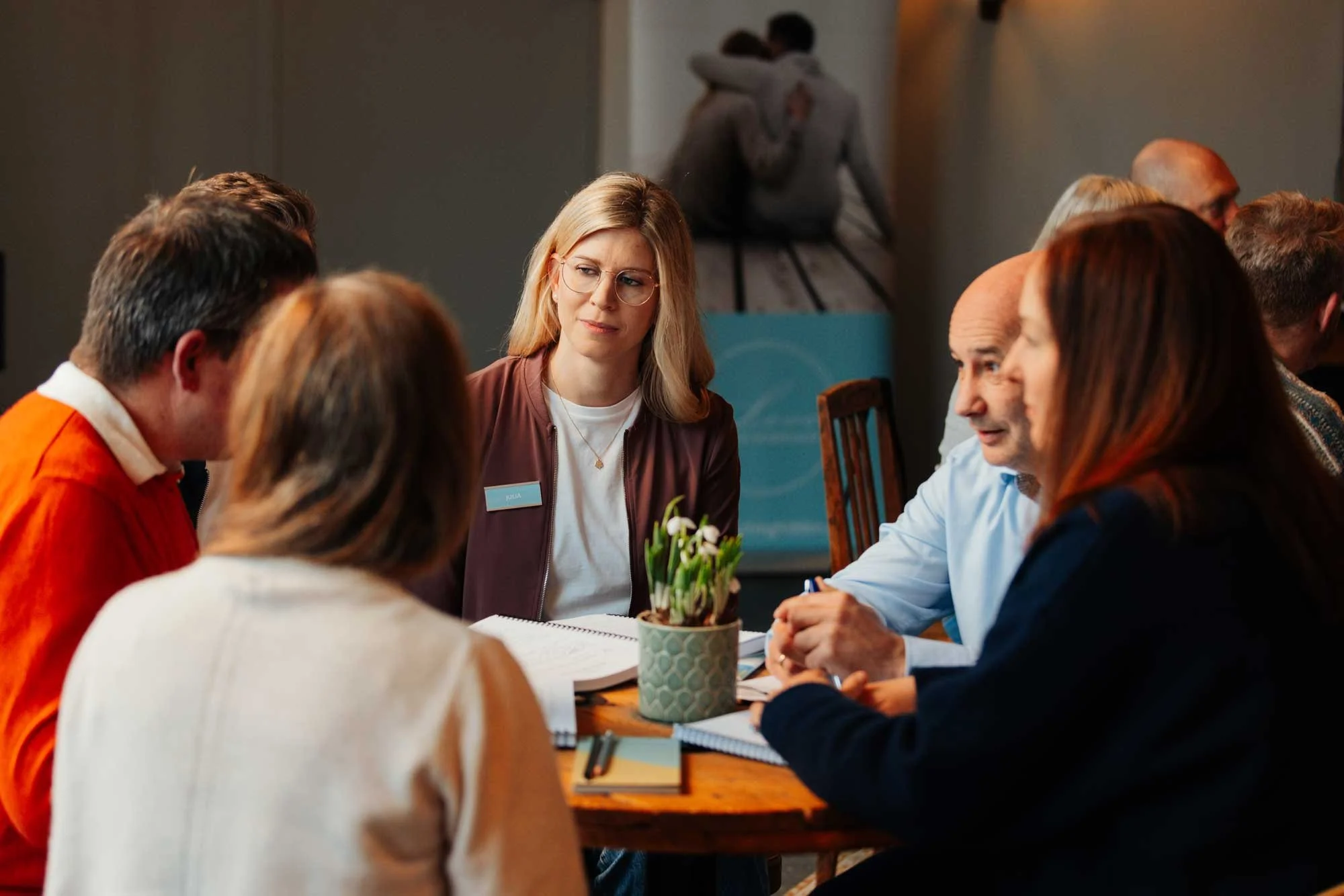 Menschen sitzen um einen Tisch bei einem Meeting, im Hintergrund eine Frau mit einem Namensschild, die zuhört, während sich hinter ihr zwei Personen umarmen.