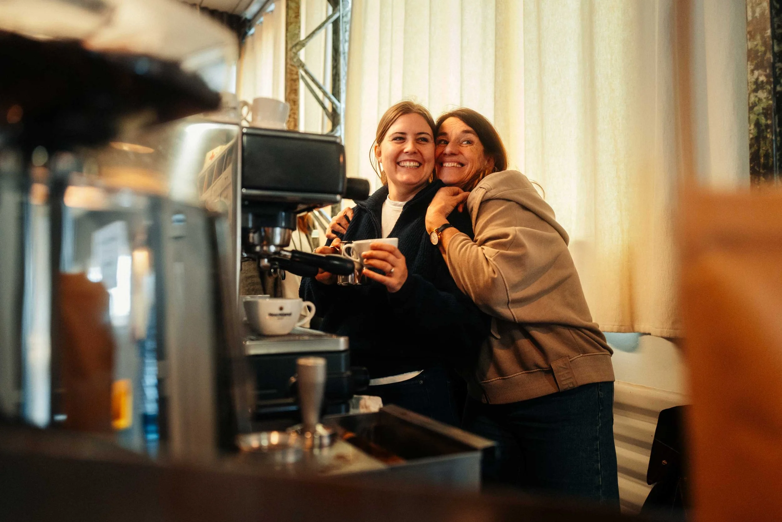 Zwei Frauen, eine jüngere und eine ältere, umarmen sich lachend in einem Café, während sie Kaffee trinken, im Hintergrund helle Vorhänge.