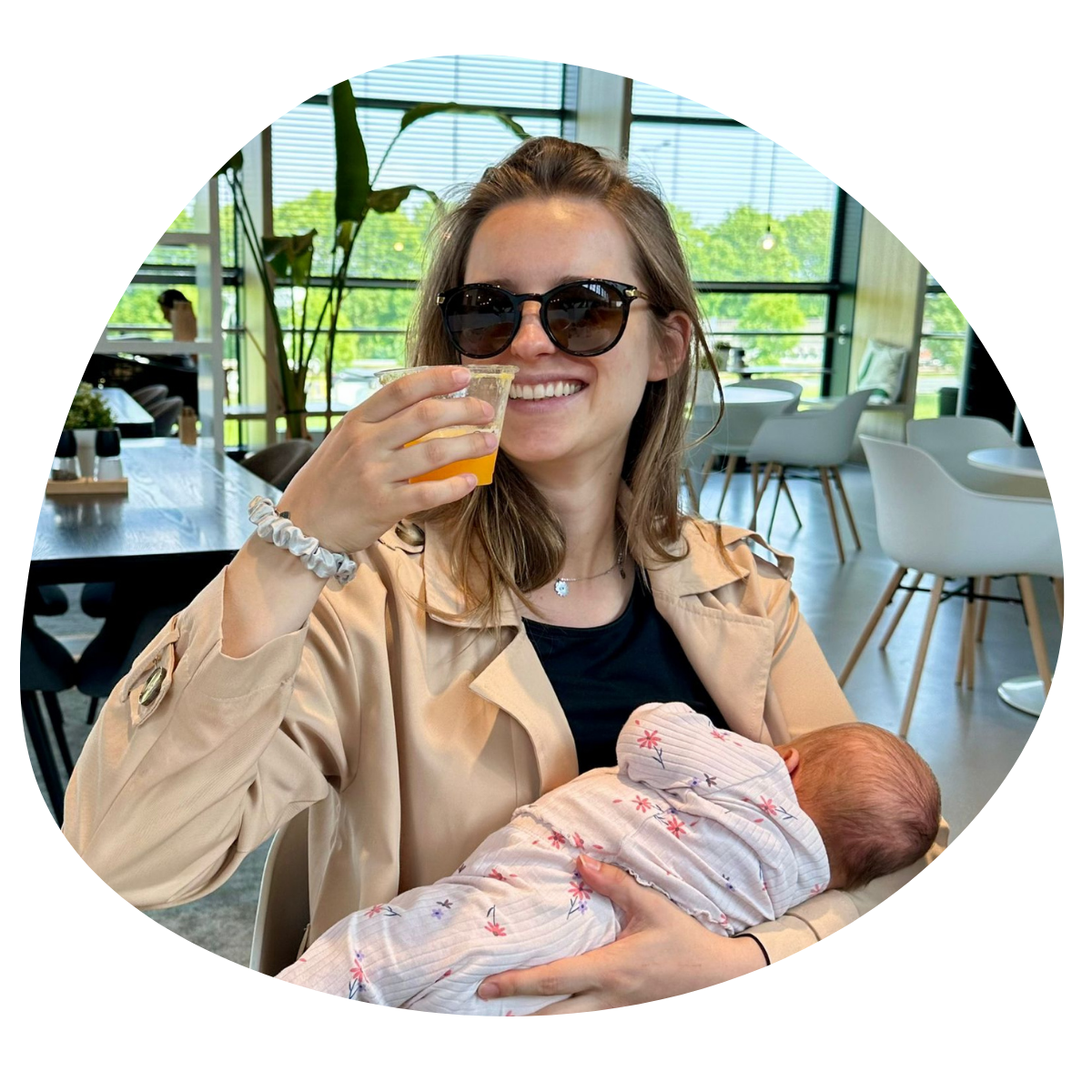 Vrouw met zonnebril die een glas limonade vasthoudt en een pasgeboren baby vasthoudt in een lichte binnenruimte met grote ramen en planten.
