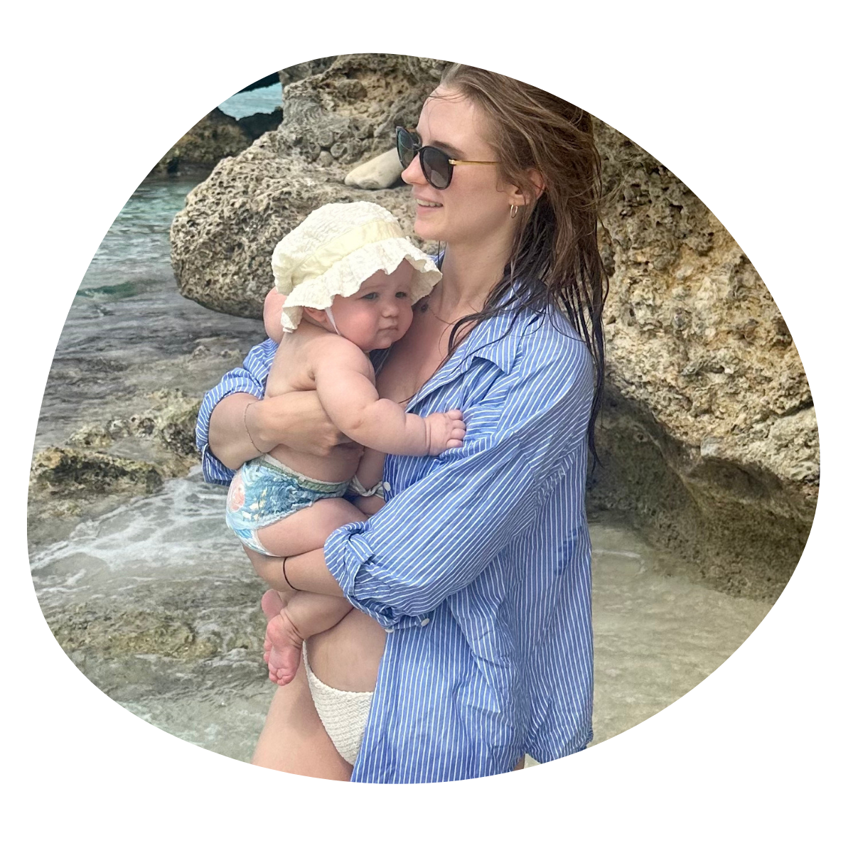 Vrouw met een baby op het strand, ze staat voor rotsen, de vrouw draagt een blauwe gestreepte blouse en zonnebril, de baby heeft een hoed en een luier.