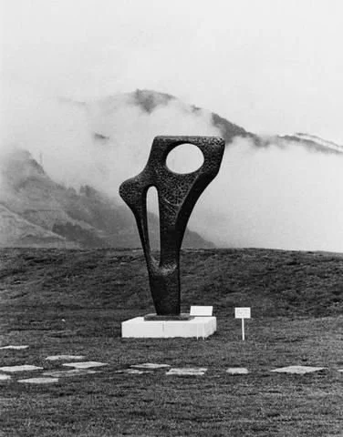 Large abstract sculpture outdoors on a grassy field with mountains and clouds in the background.