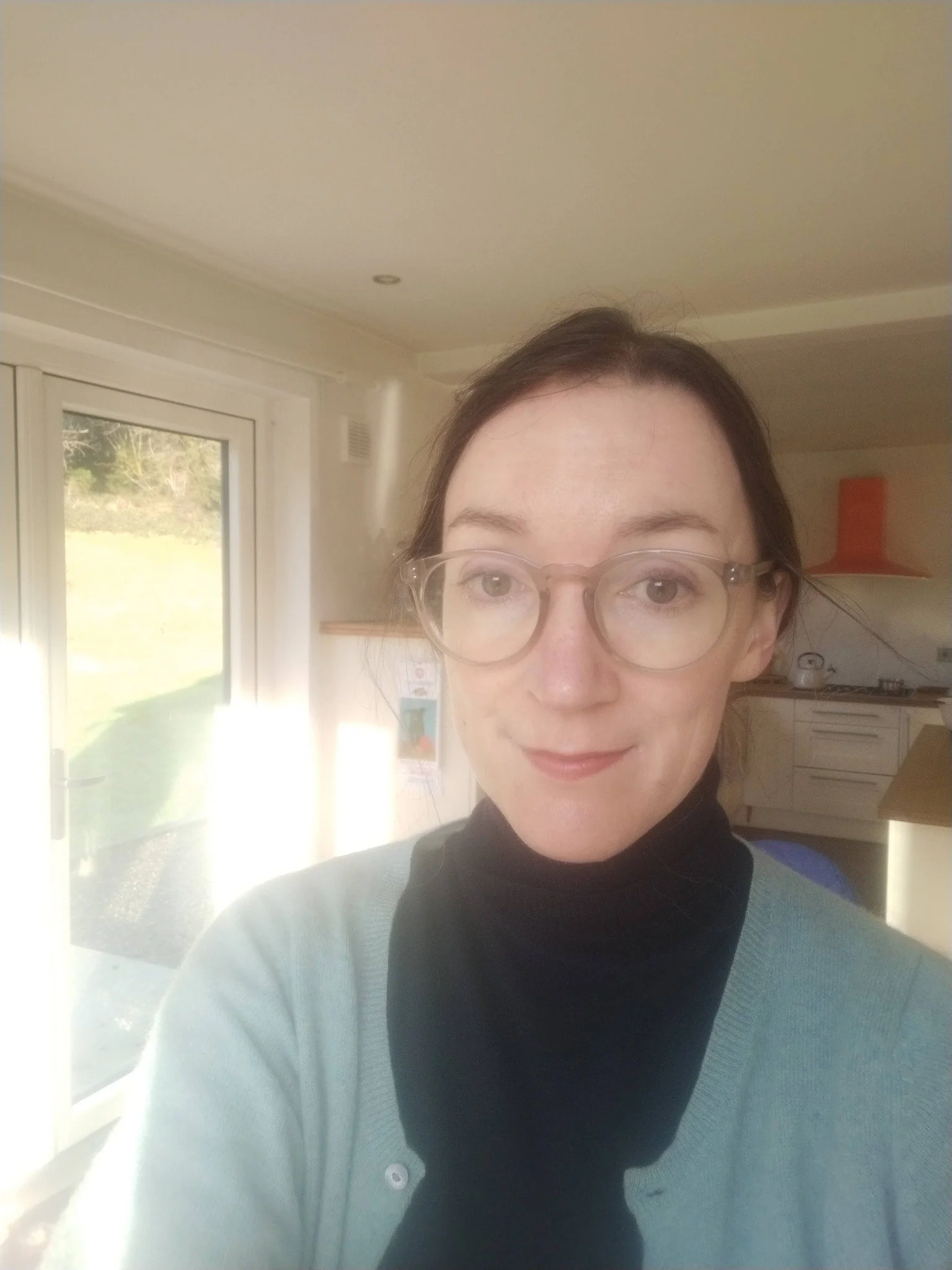 A woman with glasses and dark hair, wearing a light blue sweater and a black turtleneck, standing inside near a large window with sunlight streaming in.