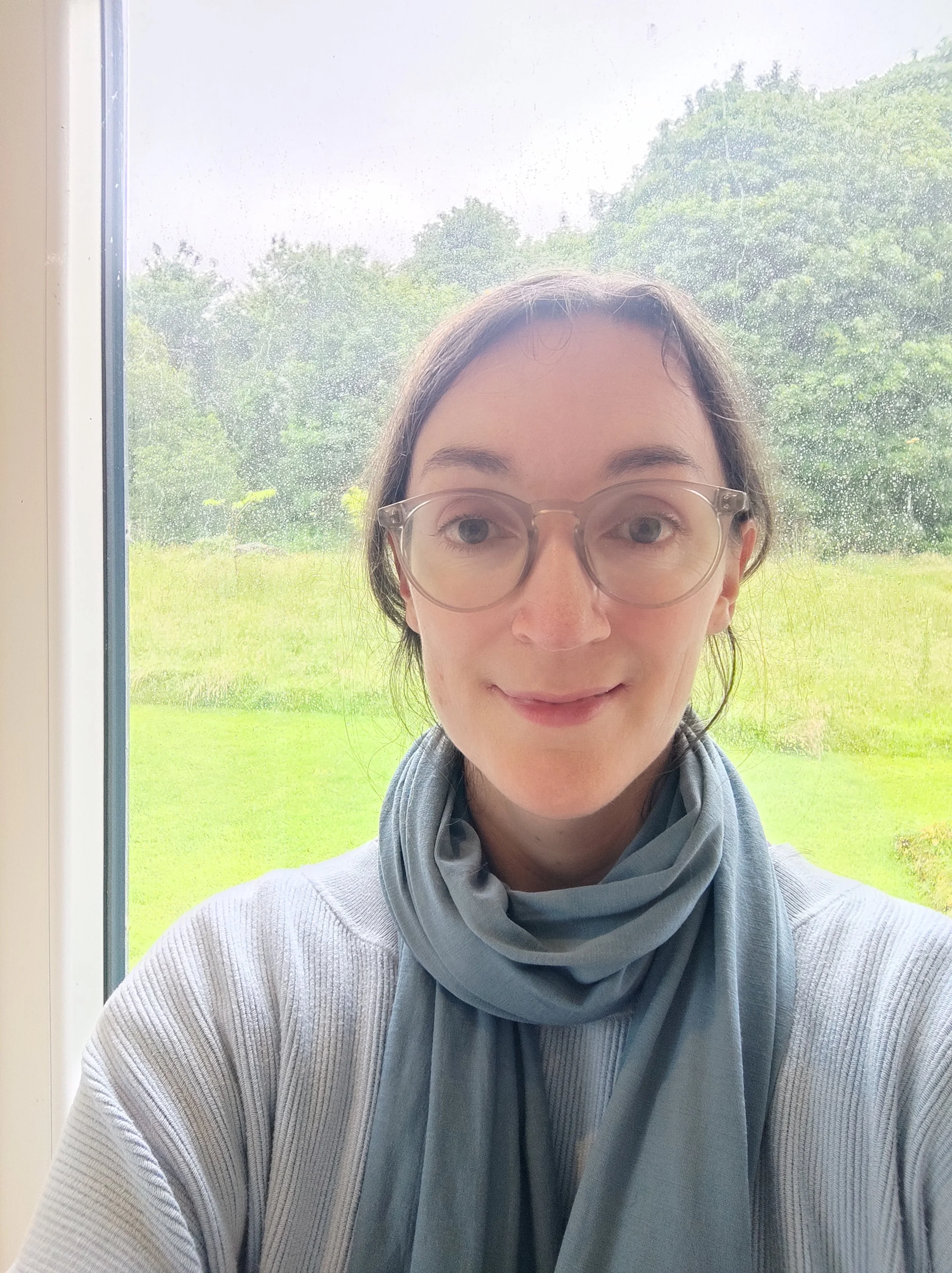 A woman with light skin and glasses is taking a selfie in front of a window. Outside, there is a green landscape with trees and grass, and it appears to be raining.