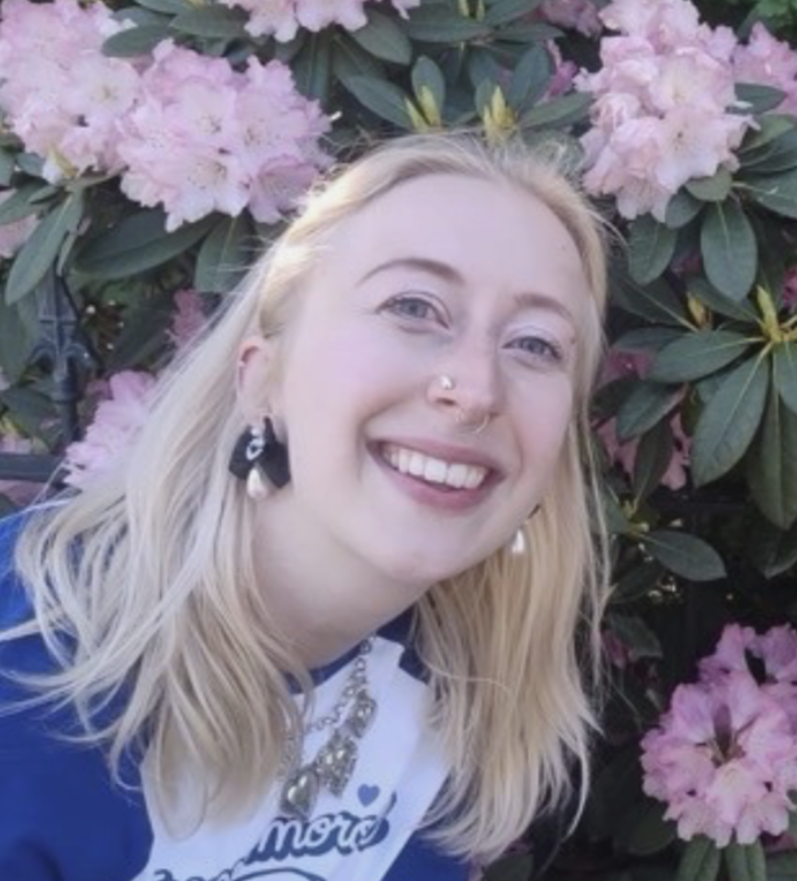 A young woman with blonde hair and piercing smiles, standing in front of pink rhododendron flowers in bloom.
