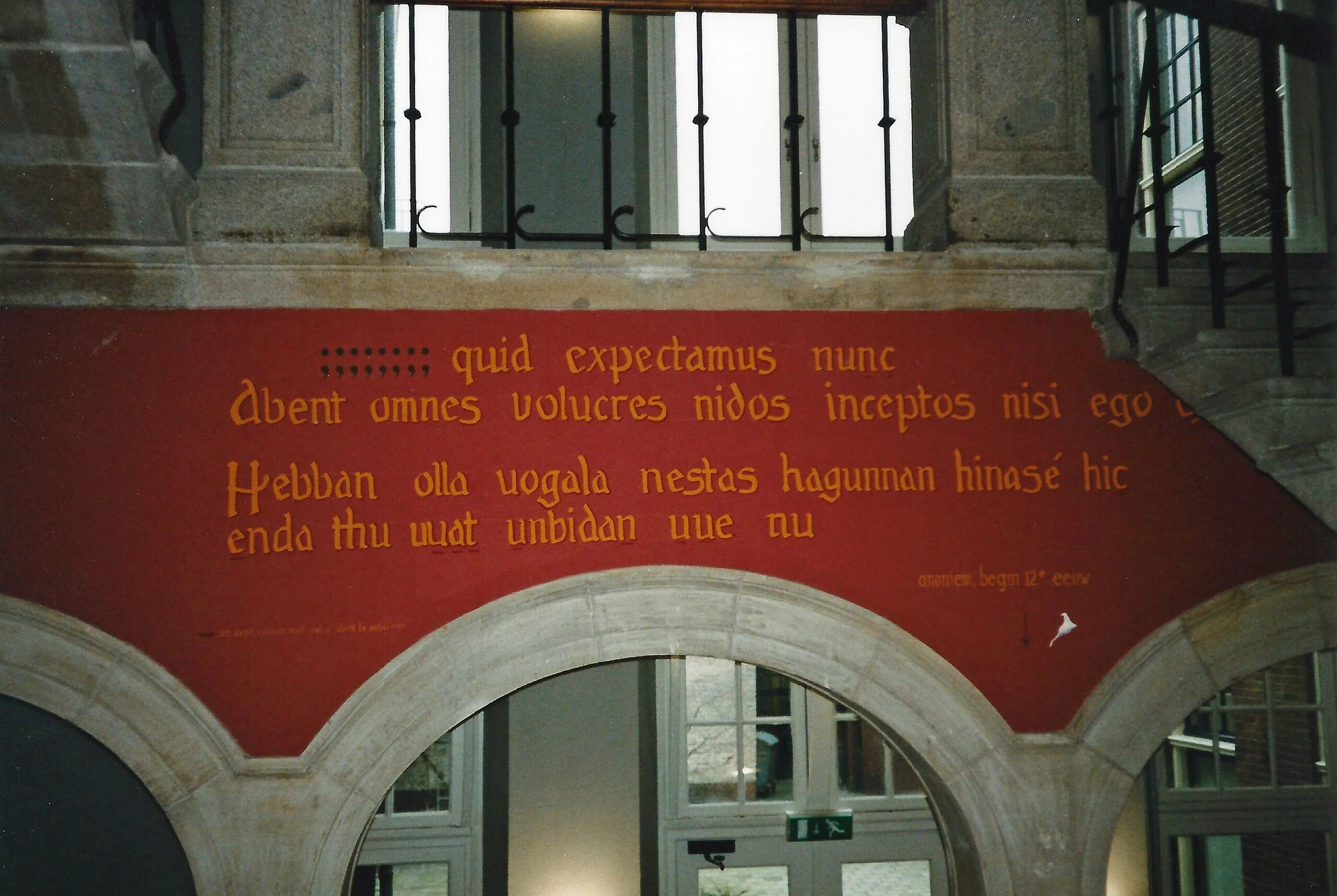 Red wall with Latin inscription in yellow lettering inside a historic building, featuring stone archways and a metal staircase.