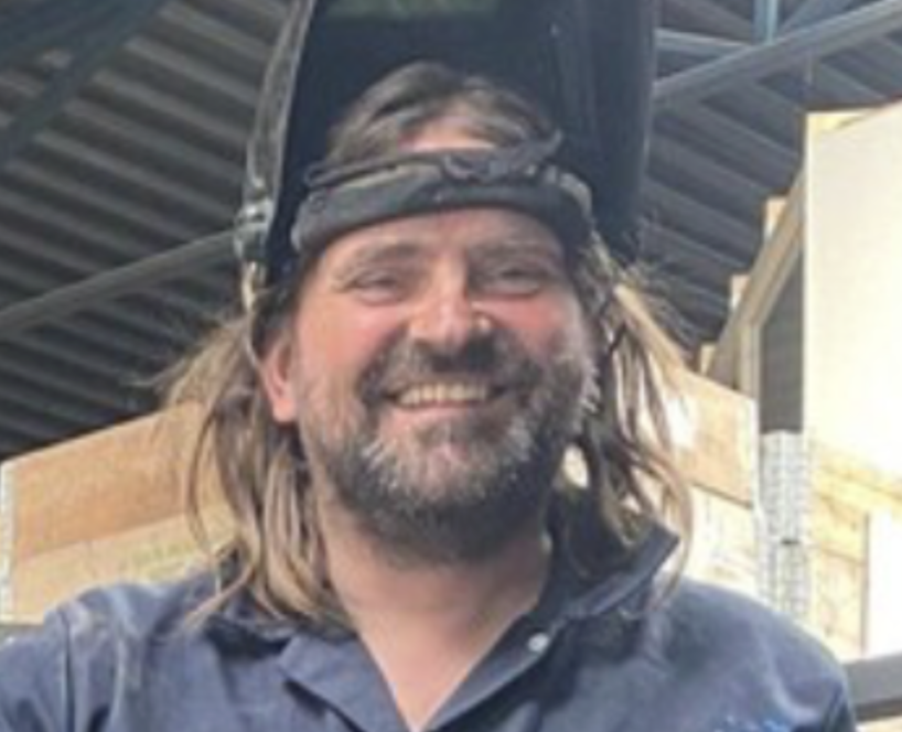 A man wearing safety goggles and a welding helmet pushed up on his head, smiling, with long hair and beard, in an industrial setting.