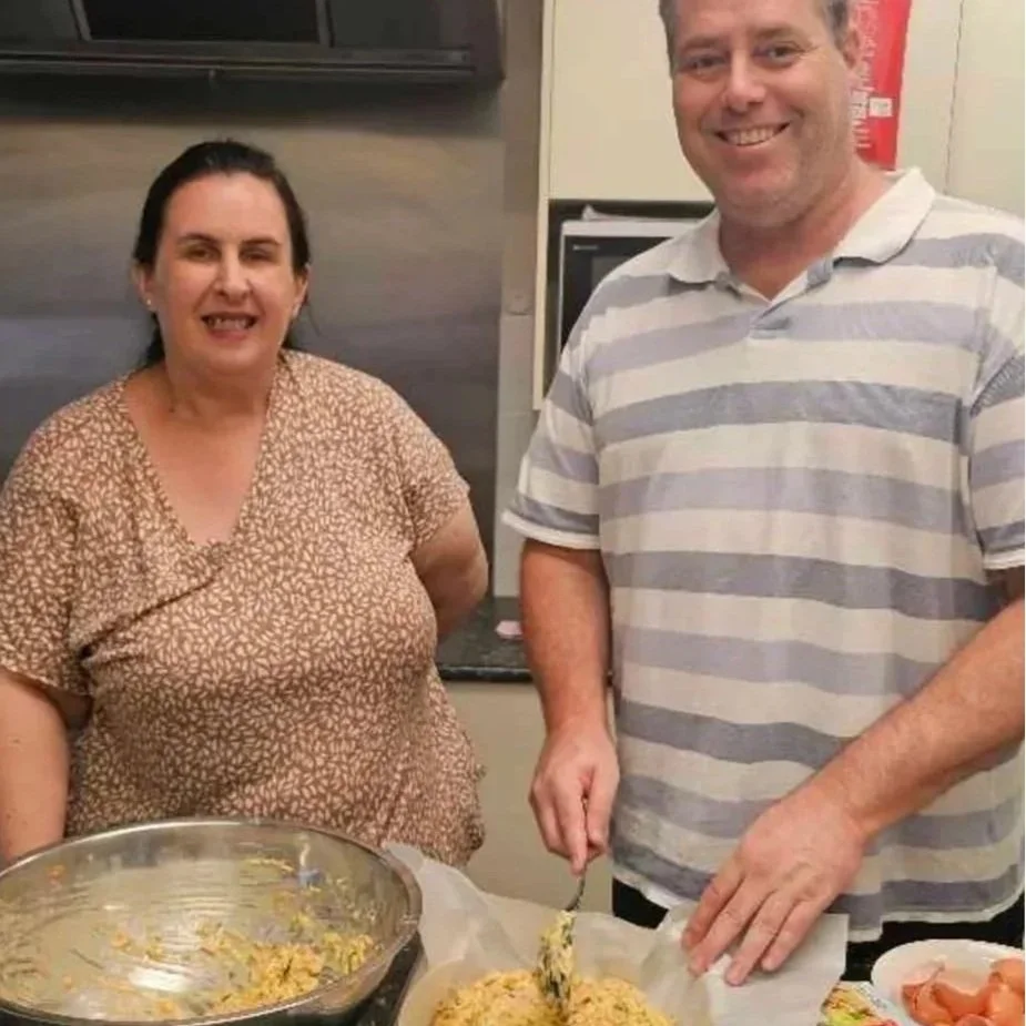 A woman and a man standing in a kitchen, smiling. The woman is wearing a patterned shirt, and the man is in a striped polo. There is a large bowl of rice or similar dish, and the man is holding a spoonful of it.