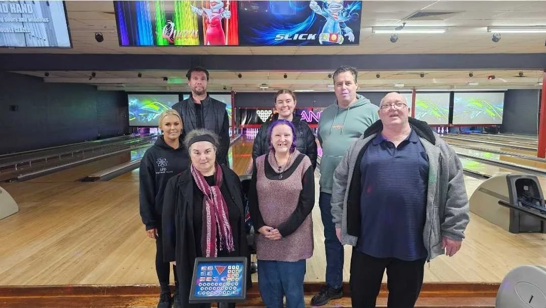 Group of seven people standing on a bowling alley with wooden lanes and digital screens above, some smiling and some looking at the camera.