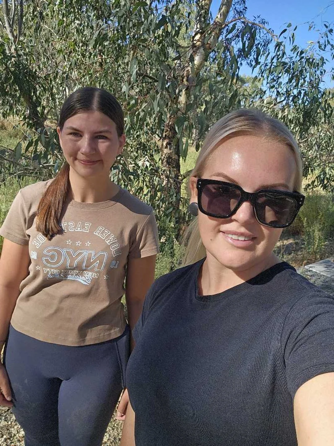 Two women taking a selfie outdoors with trees and blue sky in the background.