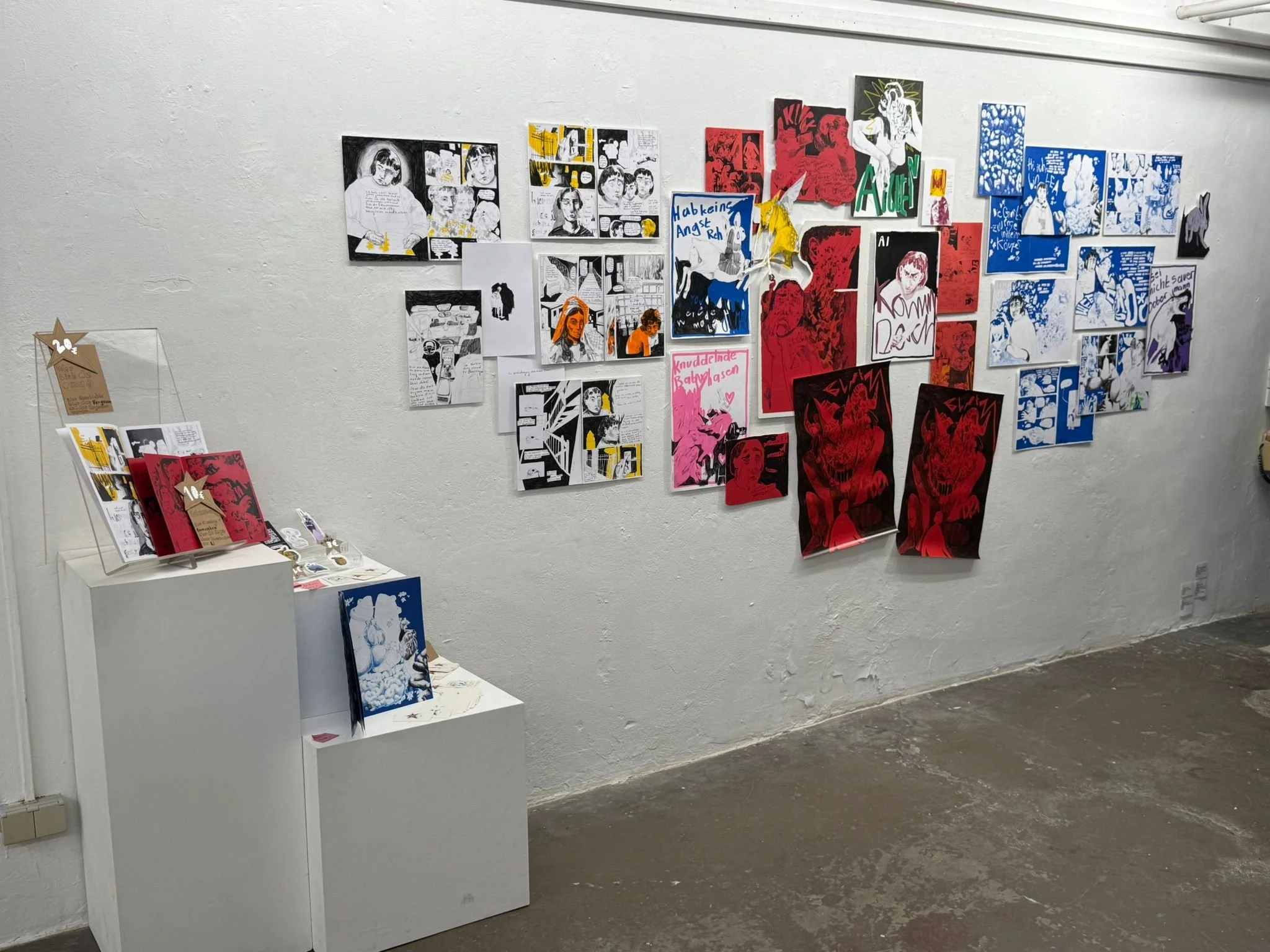 Kunstinstallation an einer weißen Wand mit bunten Comic- und lebendig wirkenden Kunstwerken, ergänzt durch Ansichts Hefte, Sticker und Visitenkarten auf weißen Ständern