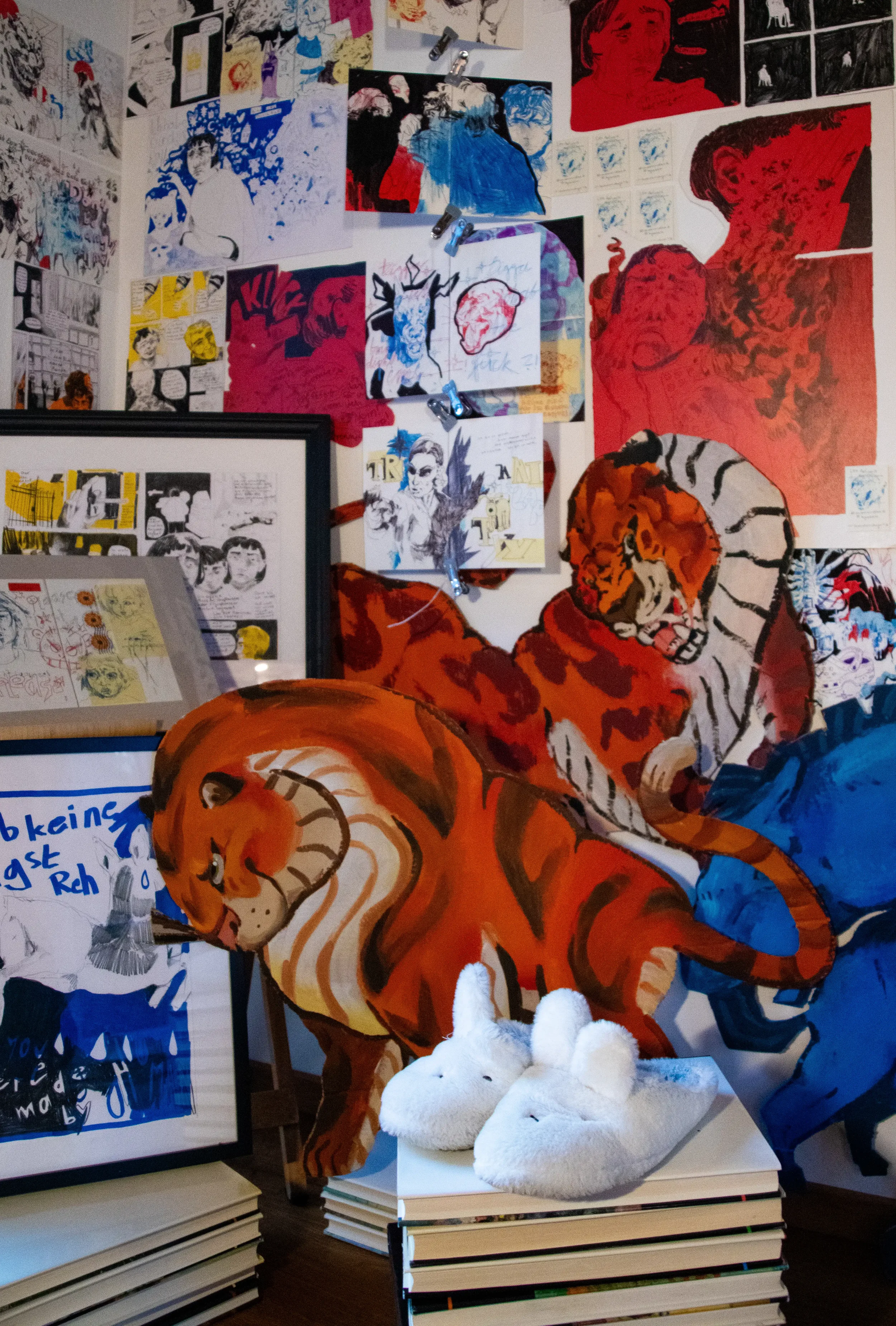 Kunstinstallation mit mehreren Gemälden, Zeichnungen, Postern und Skulpturen. Im Vordergrund zwei weiße Plüschtierhasen, darunter ein Stapel Bücher. Die Wand im Hintergrund ist bedeckt mit verschiedenen Kunstwerken, hauptsächlich in Rot- und Blautönen, darunter zwei große gemalte Tiger.