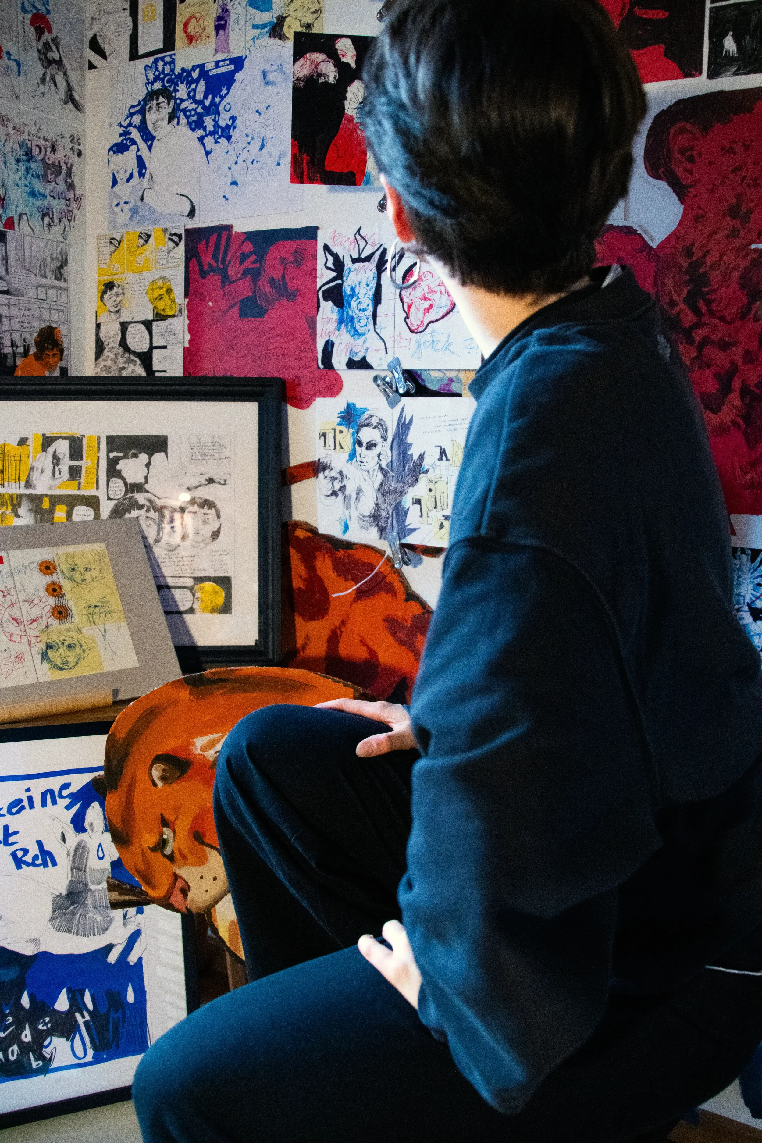Eine Person betrachtet eine Wand voller verschiedener Kunstwerke, darunter Gemälde, Skizzen und Comics in bunten Farben.