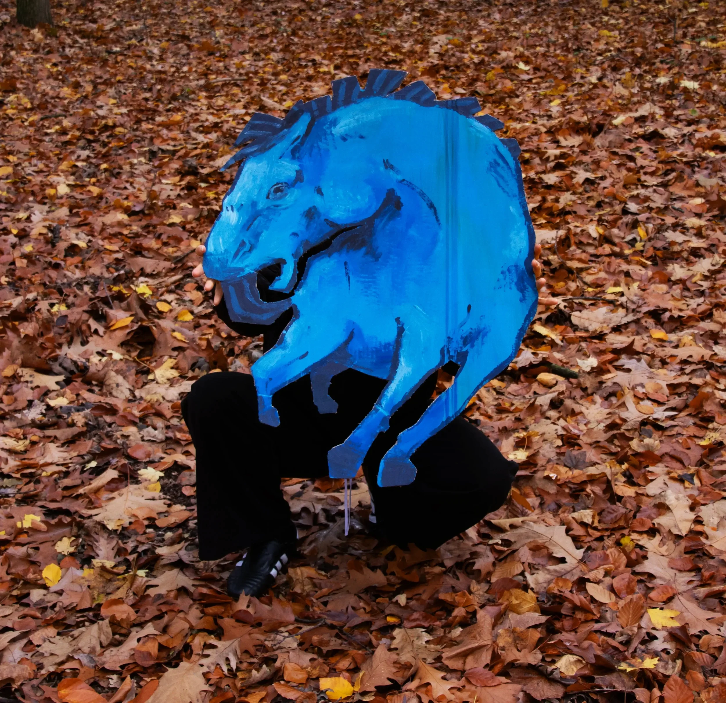 Person hält ein Kunstwerk in Form eines blauen Pferdekopfes in einem herbstlichen Wald mit Laub bedecktem Boden