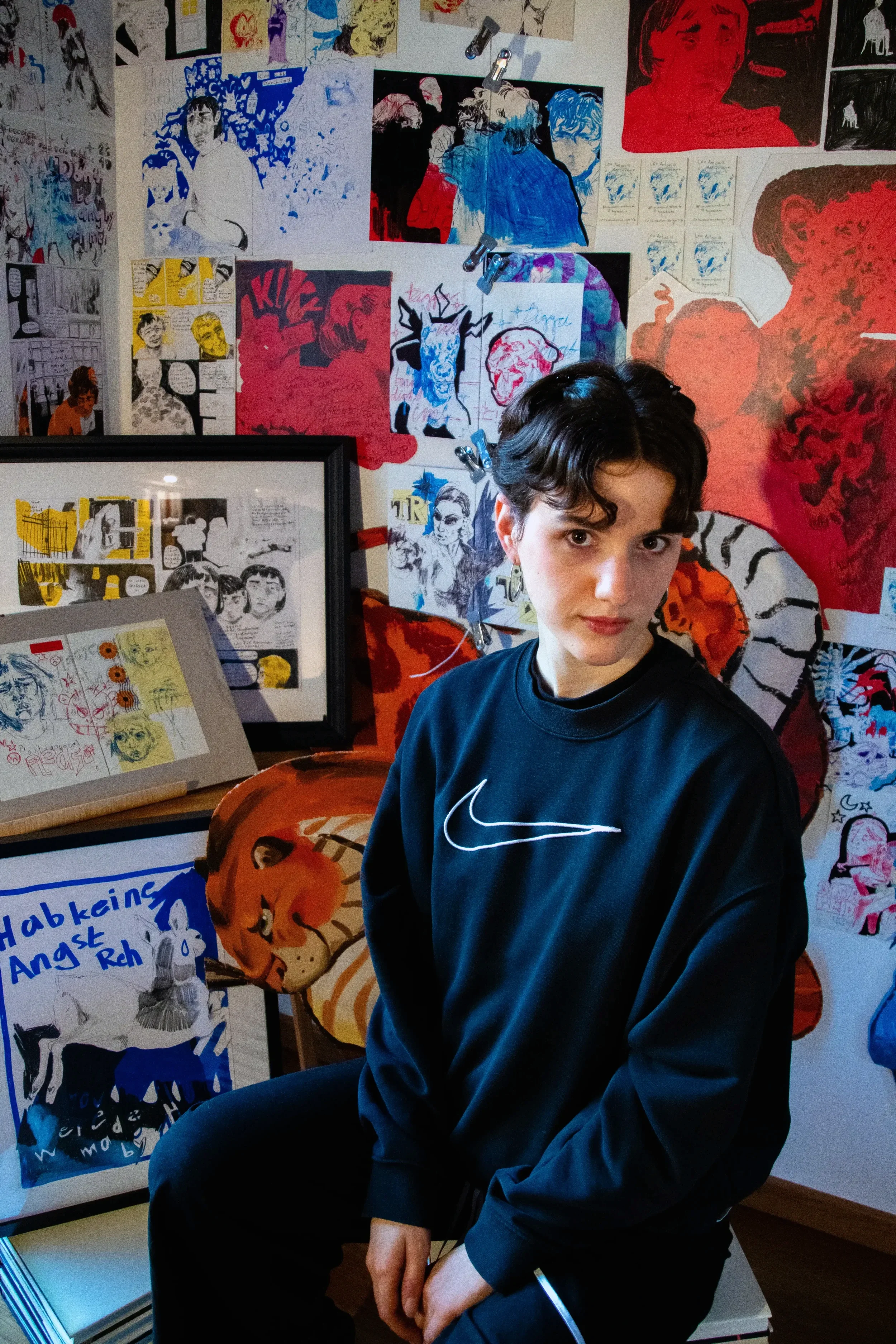 Junge Frau mit dunklem, kurz gewelltem Haar in schwarzem Nike-Sweatshirt, sitzend vor einer Wand voller bunter, künstlerischer Poster und Zeichnungen.