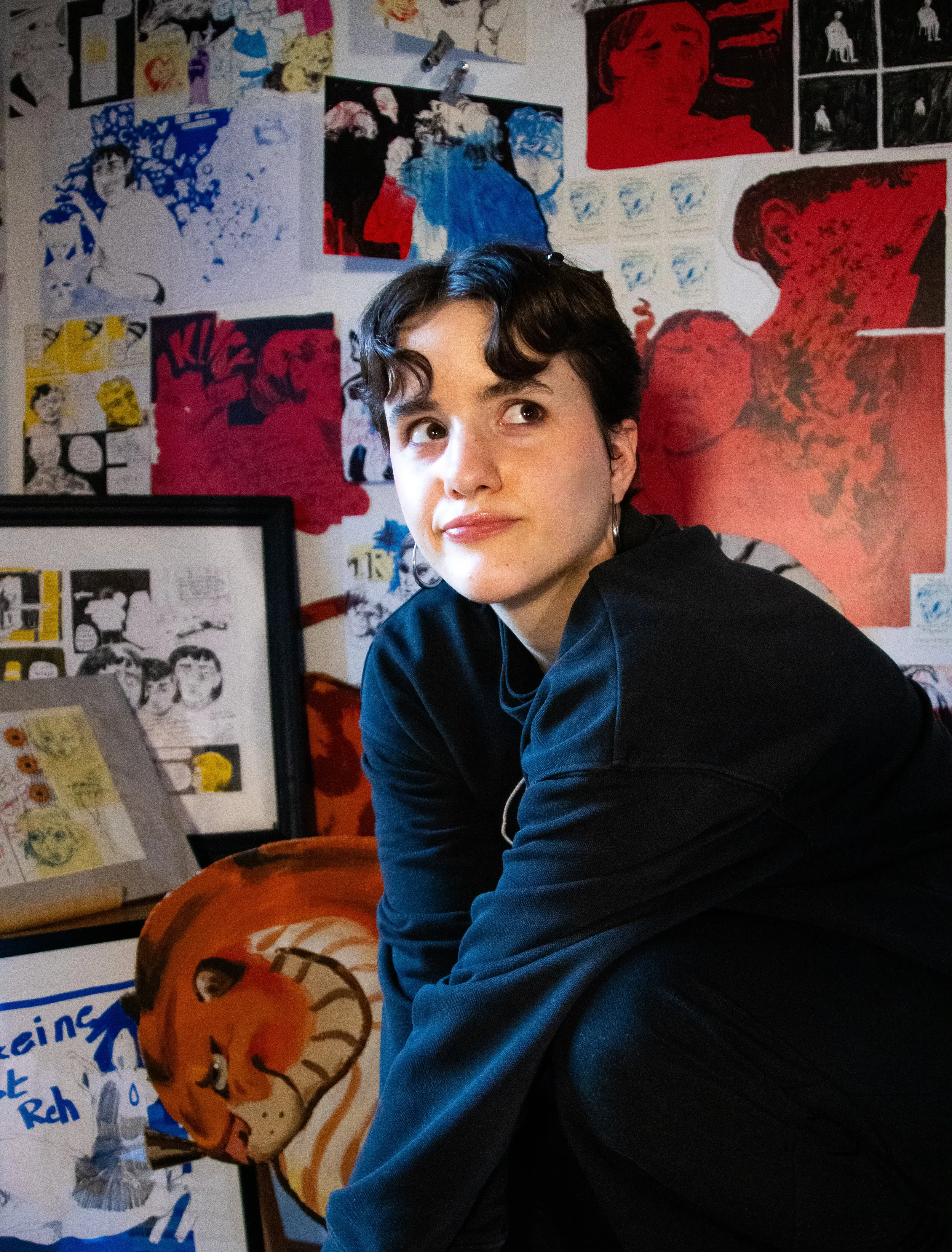 Junge Frau mit dunklen, kurzen Haaren in schwarzer Jacke, sitzt vor einer Wand voller bunter Pop-Art und Comics, mit einem großen Bild eines Tigers im Vordergrund.
