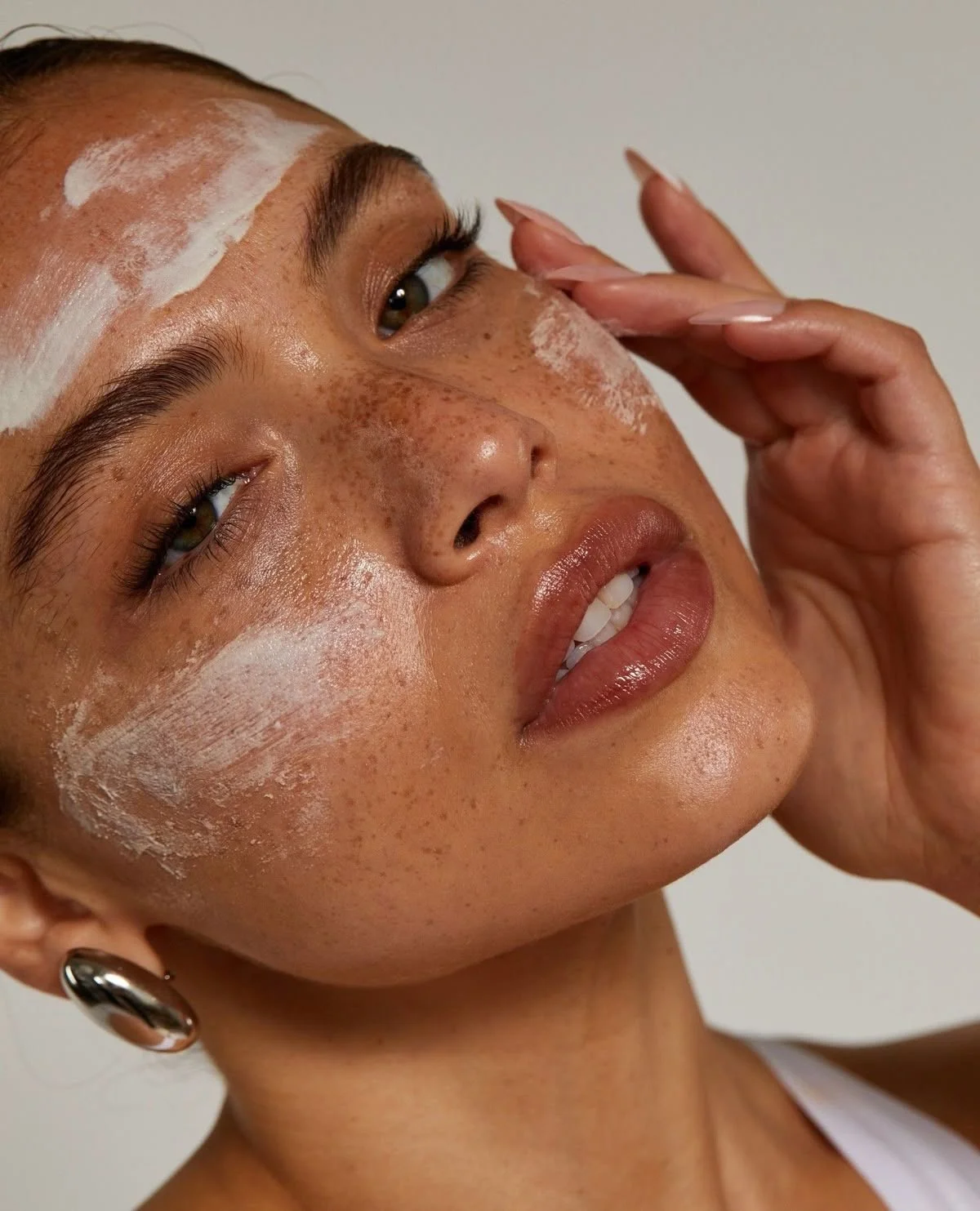 A close-up of a woman with freckles, wearing makeup, with white cream on her face, touching her face with one hand, and wearing earrings.