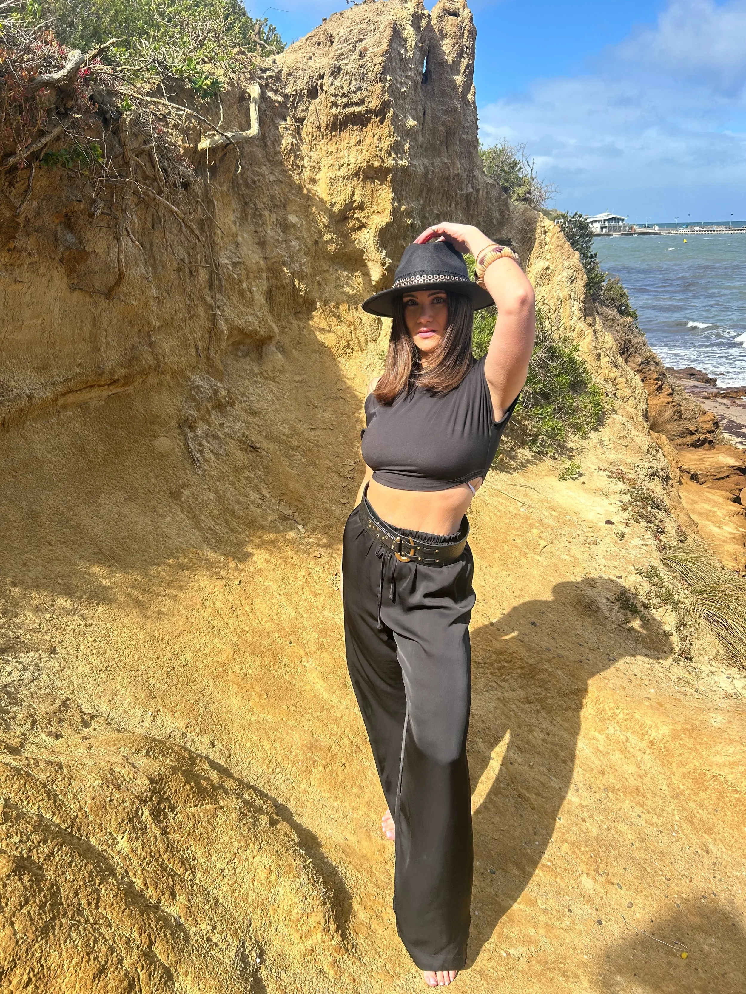 A woman wearing a black crop top, black wide-leg pants, and a black hat posing on a sandy beach with a rocky cliff and ocean in the background.