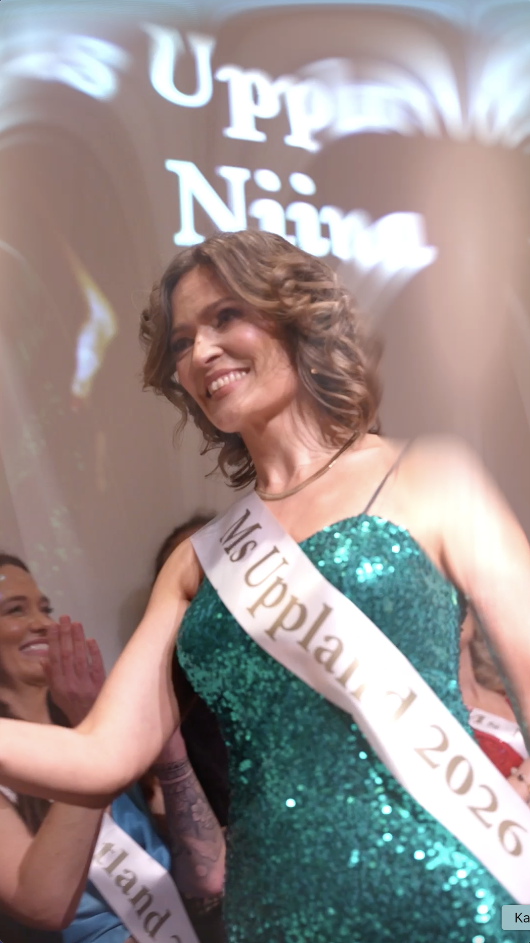 En kvinna som vunnit Miss Uppsalas 2023, klädd i en glittrande grön klänning och med en vinnarskede, ler och är glad.