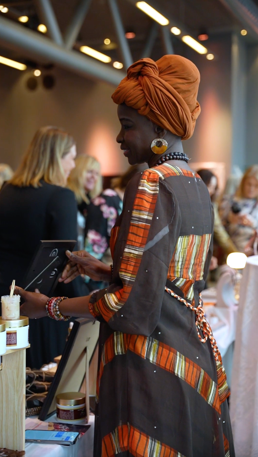 En kvinna med mörk hud och stort orange Headwrap, färgglad klädsel med orange, svart och vita ränder, står vid ett bord i ett evenemang eller butik, omgiven av människor i bakgrunden.