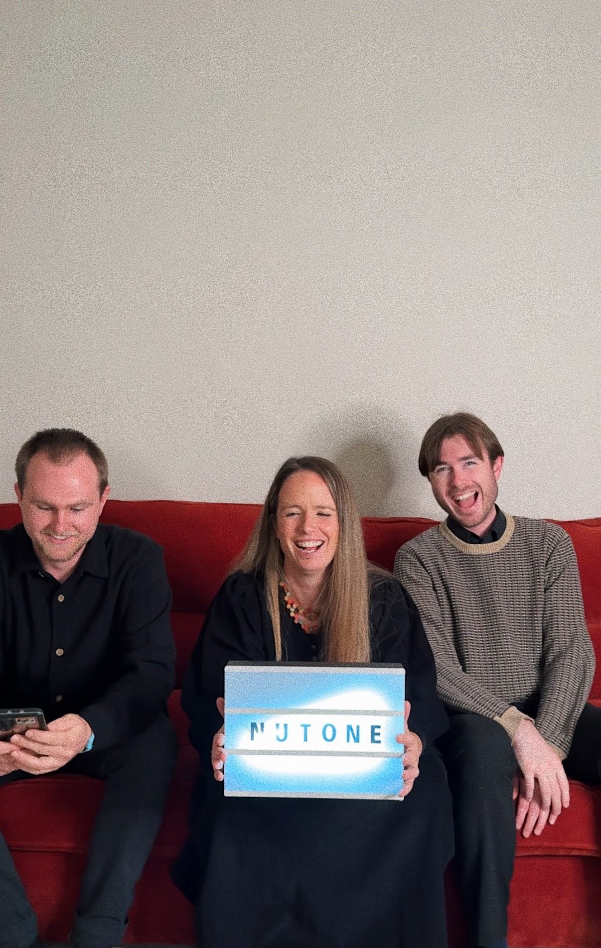 Three people sitting on a red couch, smiling and laughing. The woman in the middle is holding a lightbox sign that spells 'NUTONE'.