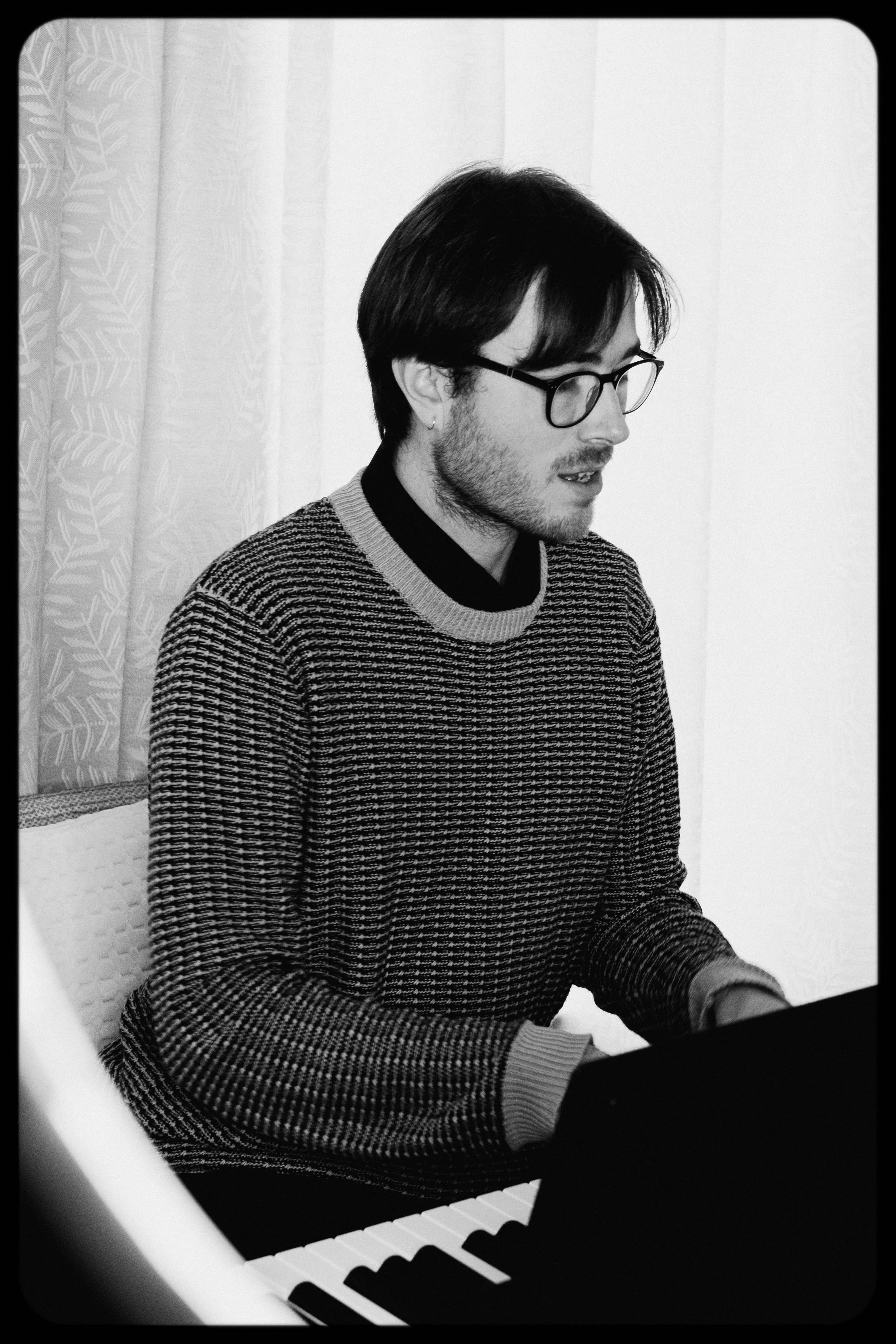 A young man with glasses and dark hair is playing a piano indoors. He is wearing a patterned sweater and appears focused on the music.