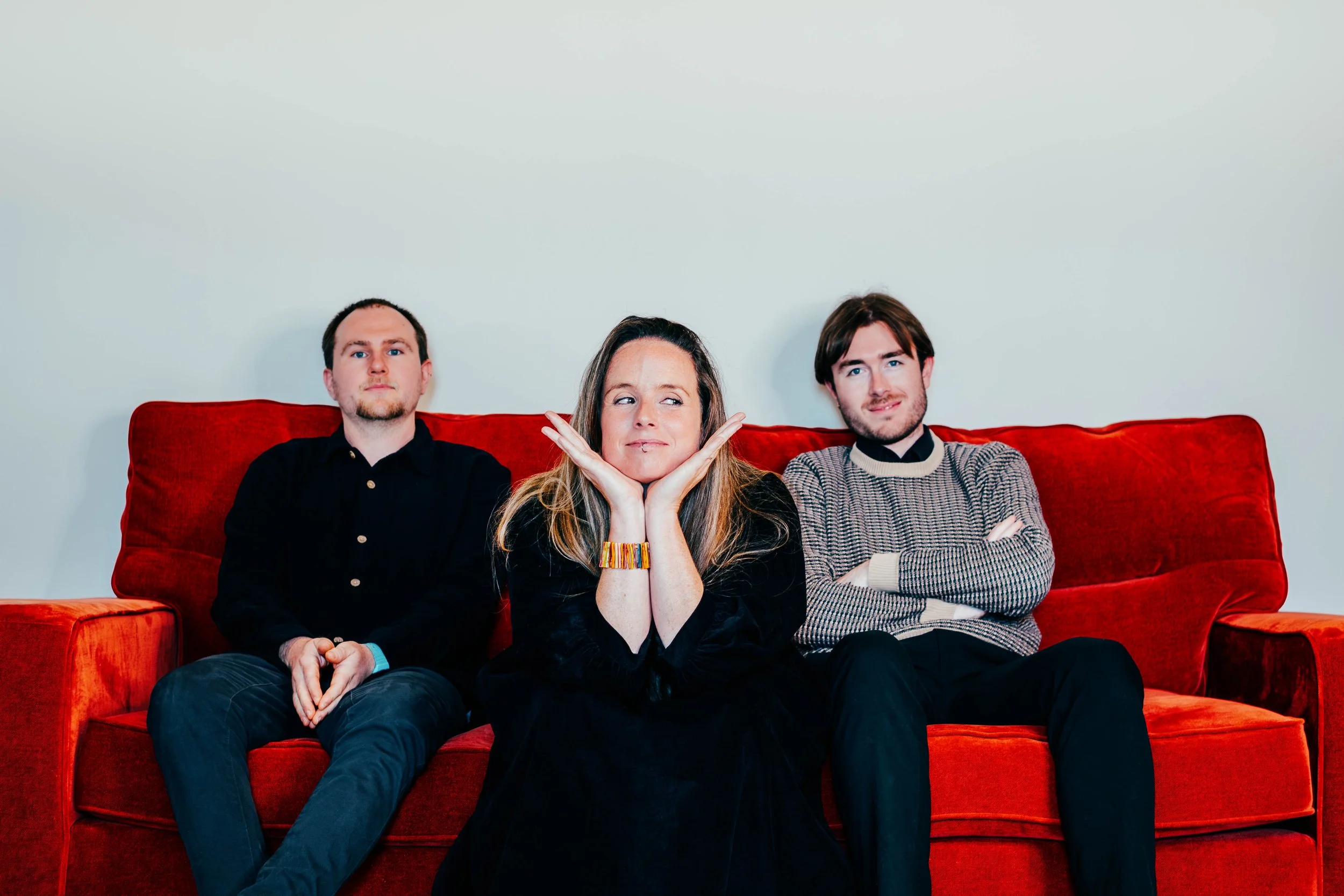 Three people sitting on a red couch against a plain white wall, with the woman in the center resting her chin on her hands and smiling, flanked by two men with arms crossed.