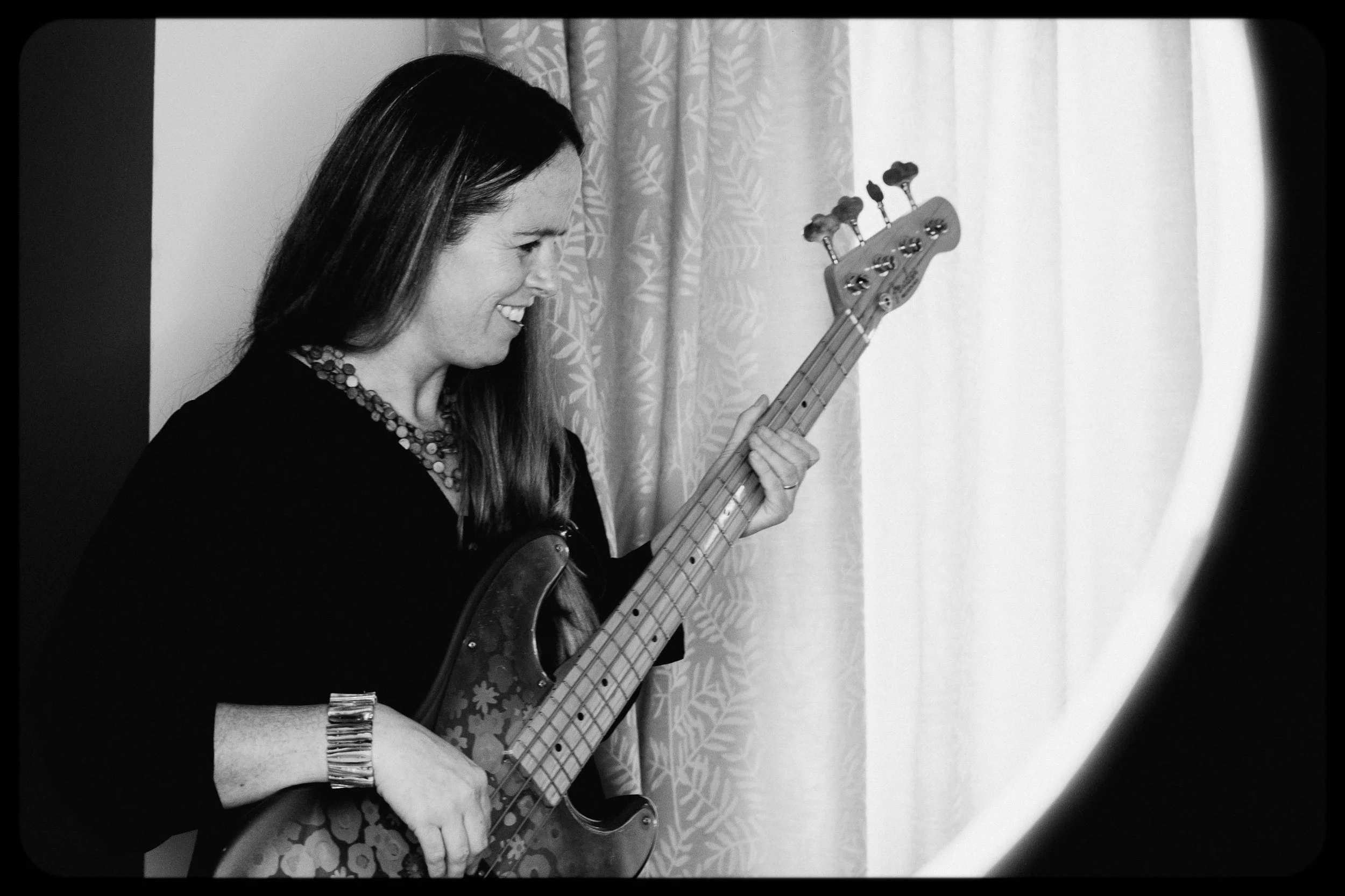 A woman smiling and playing an electric bass guitar indoors, standing next to curtains.