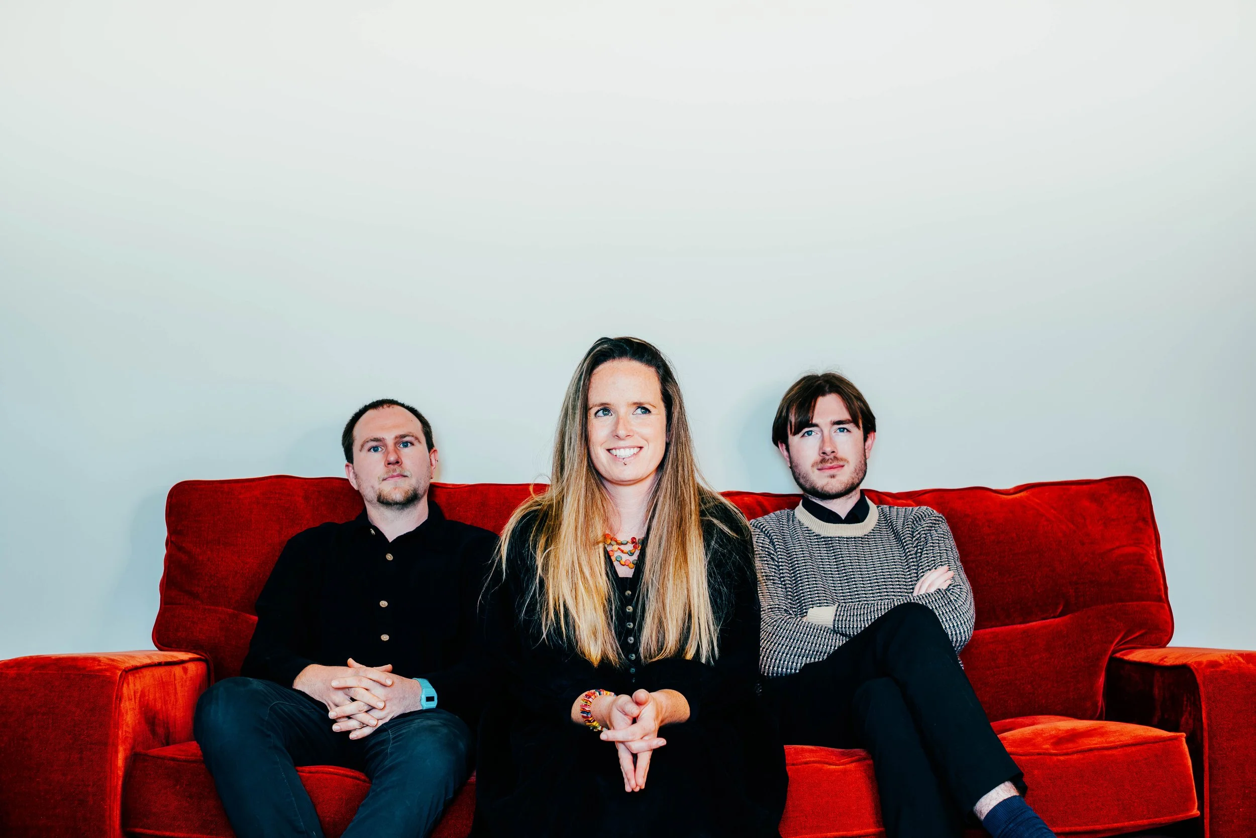 Three people sitting on a red sofa against a white background, with the woman in the middle smiling and the two men on either side looking serious.