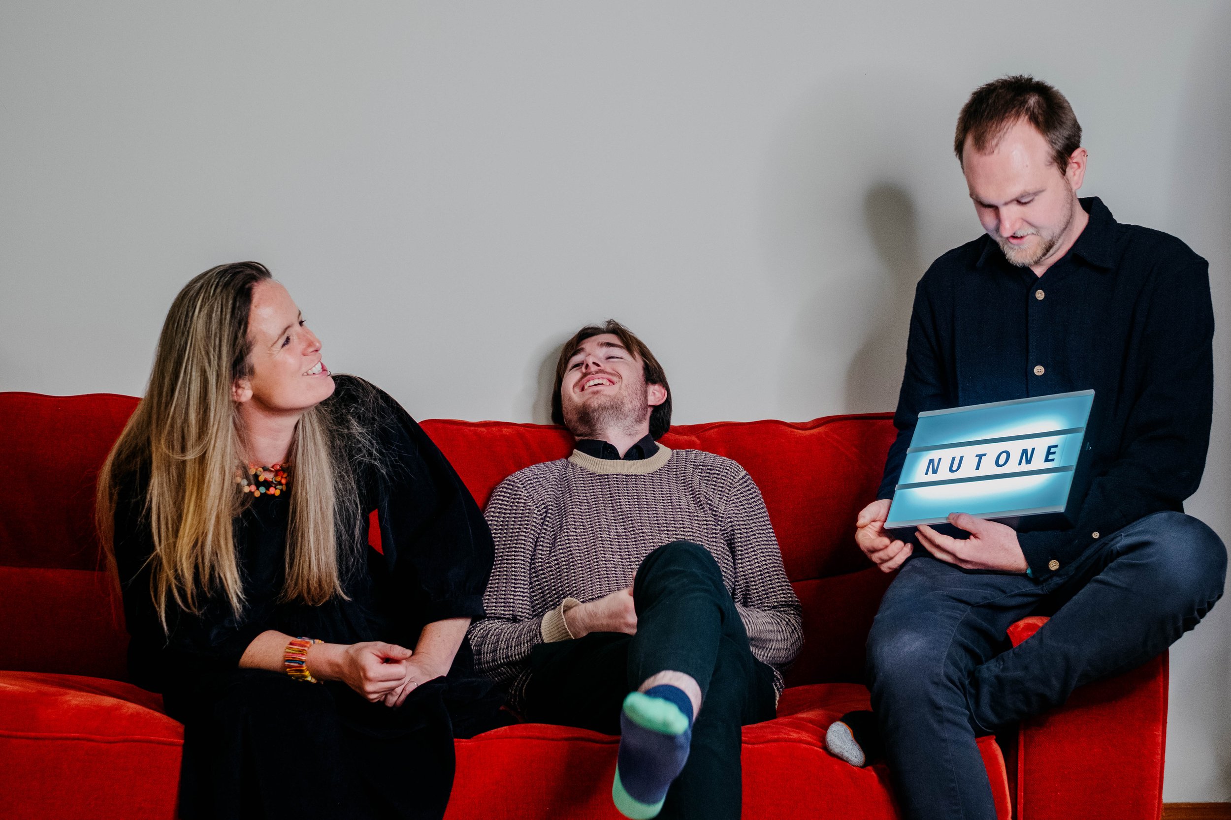 Three people sitting on a red couch, two women and one man, laughing and smiling. A man on the right is holding a tablet with a glowing screen that says 'NUTONE'. The women are looking at each other and the man is looking down at the tablet.