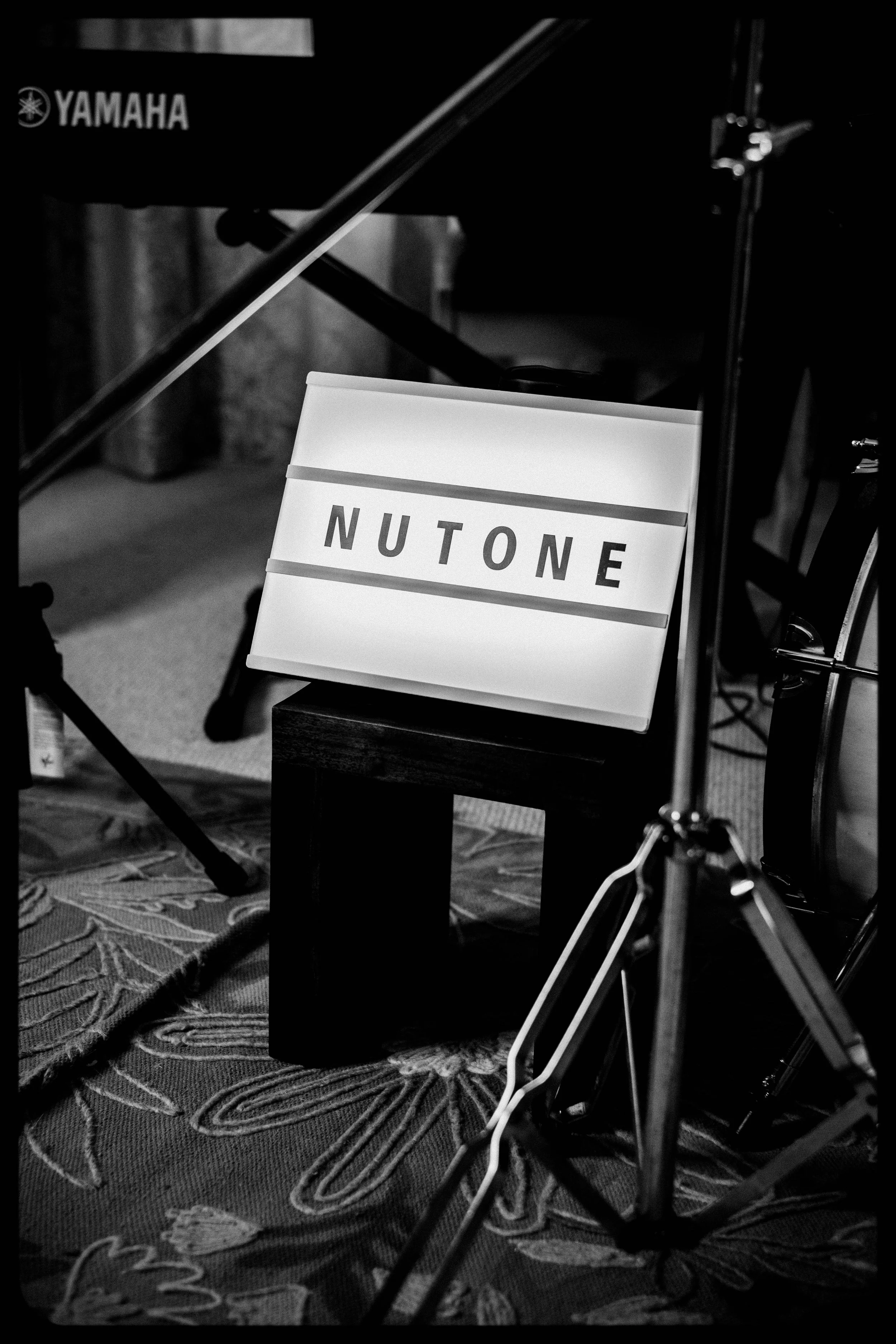 A lightbox sign with the word "NUTONE" on it, placed on a small wooden stool part of a music or recording setup, with a Yamaha keyboard and drum set partially visible in a dimly lit room.