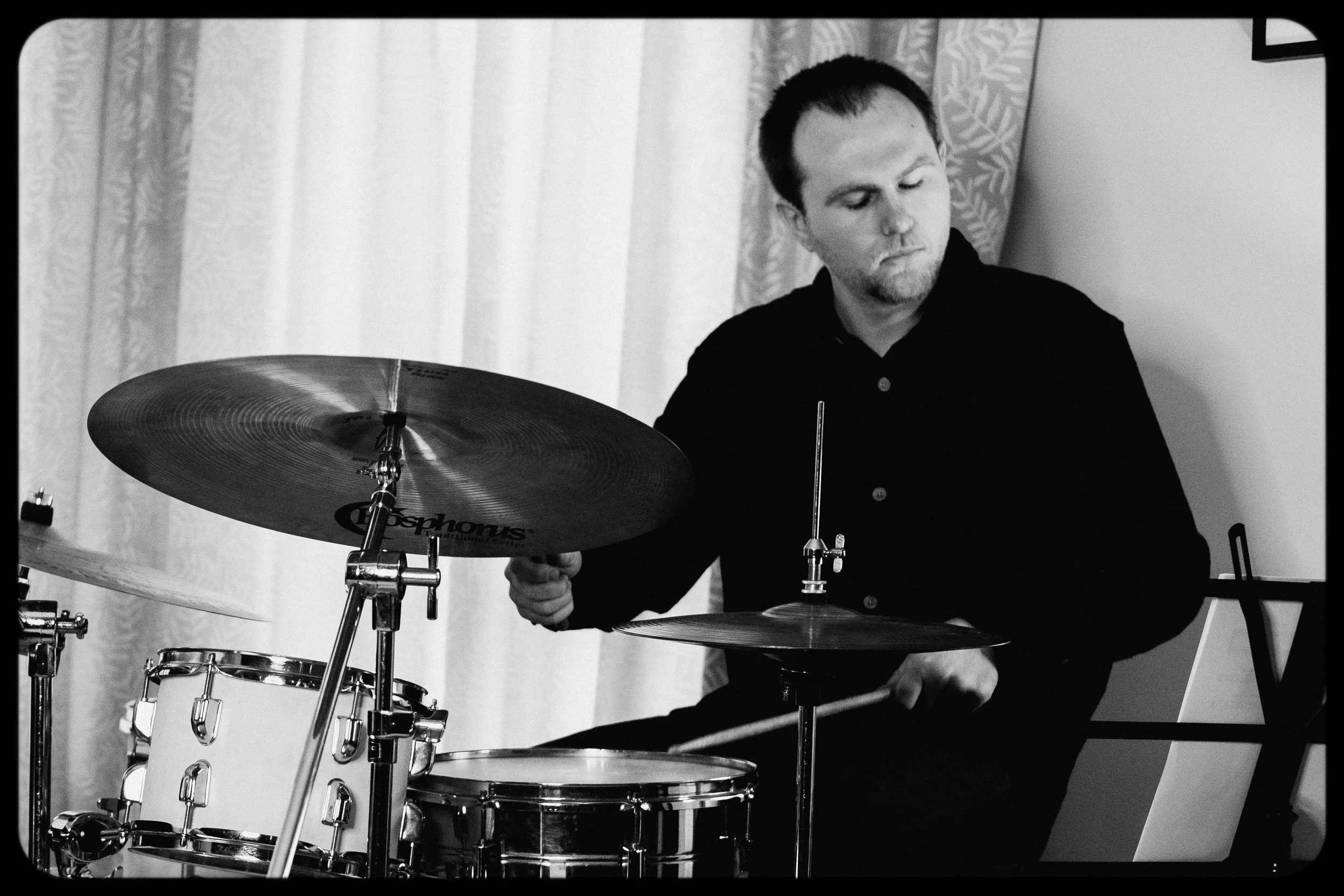 A man with short hair and a beard playing a drum set, with cymbals and a snare drum, in a room with curtains.