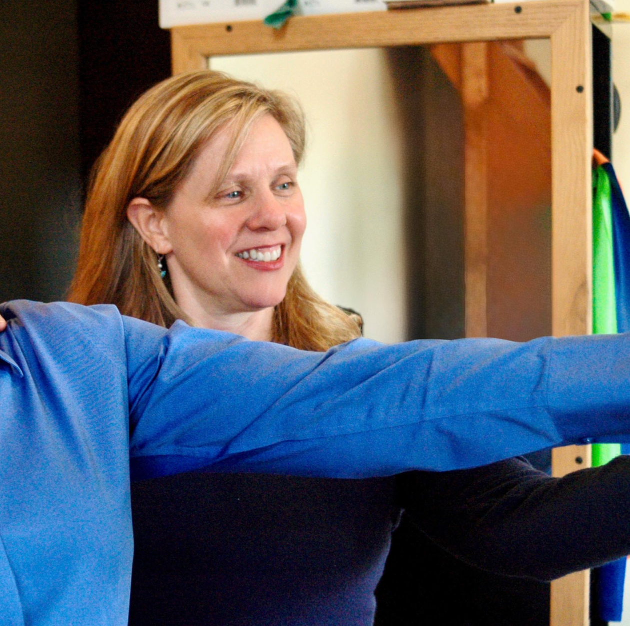 A woman with red hair smiling and pointing, with another person in a blue shirt in front of her. They appear to be in a room with a wooden structure and colorful items in the background.