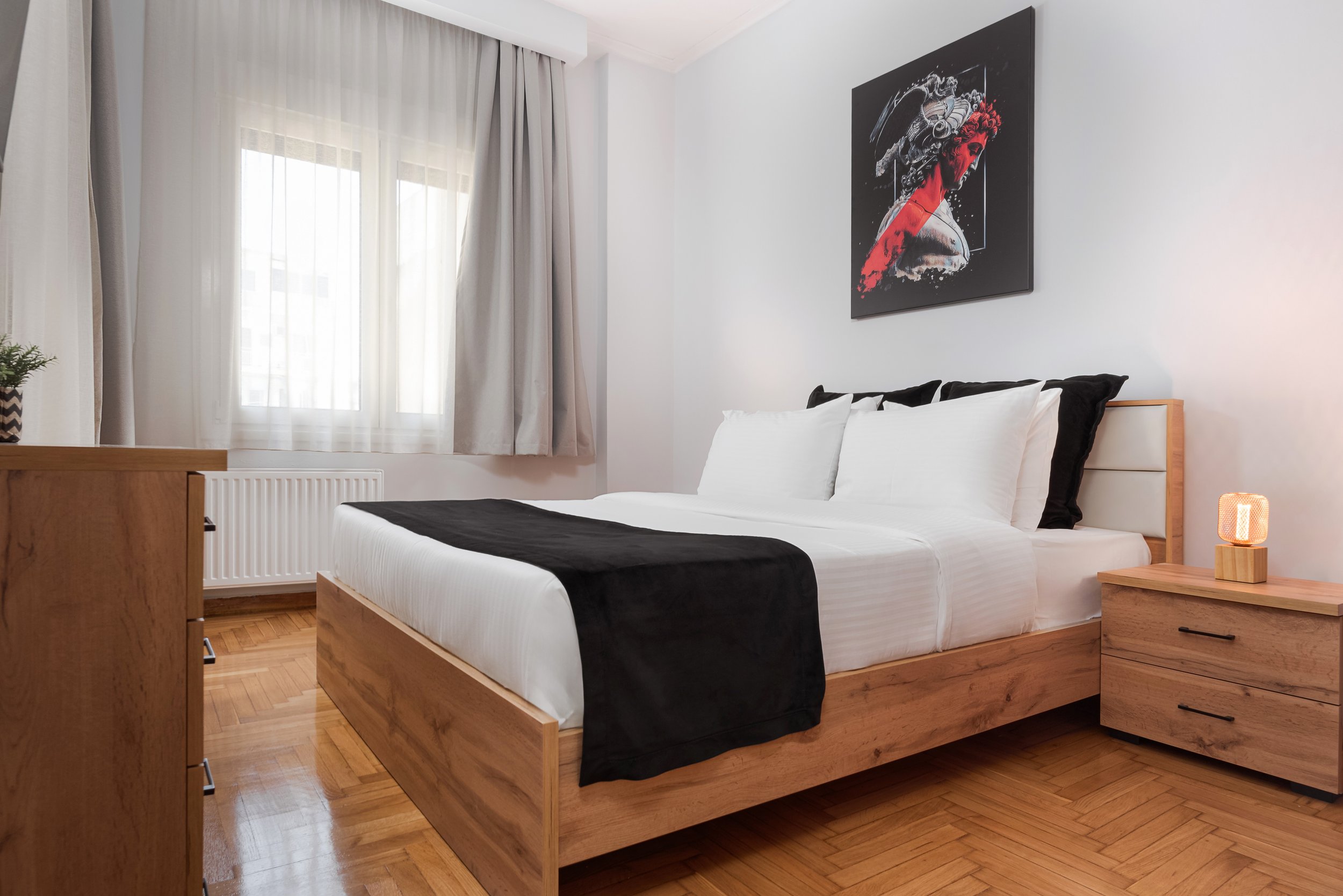 Bedroom with a wooden bed, white bedding, black accent pillows, black runner, wooden nightstand with lamp, window with sheer curtains, artwork of a woman with a red streak on her face, wooden wardrobe, and hardwood floor.