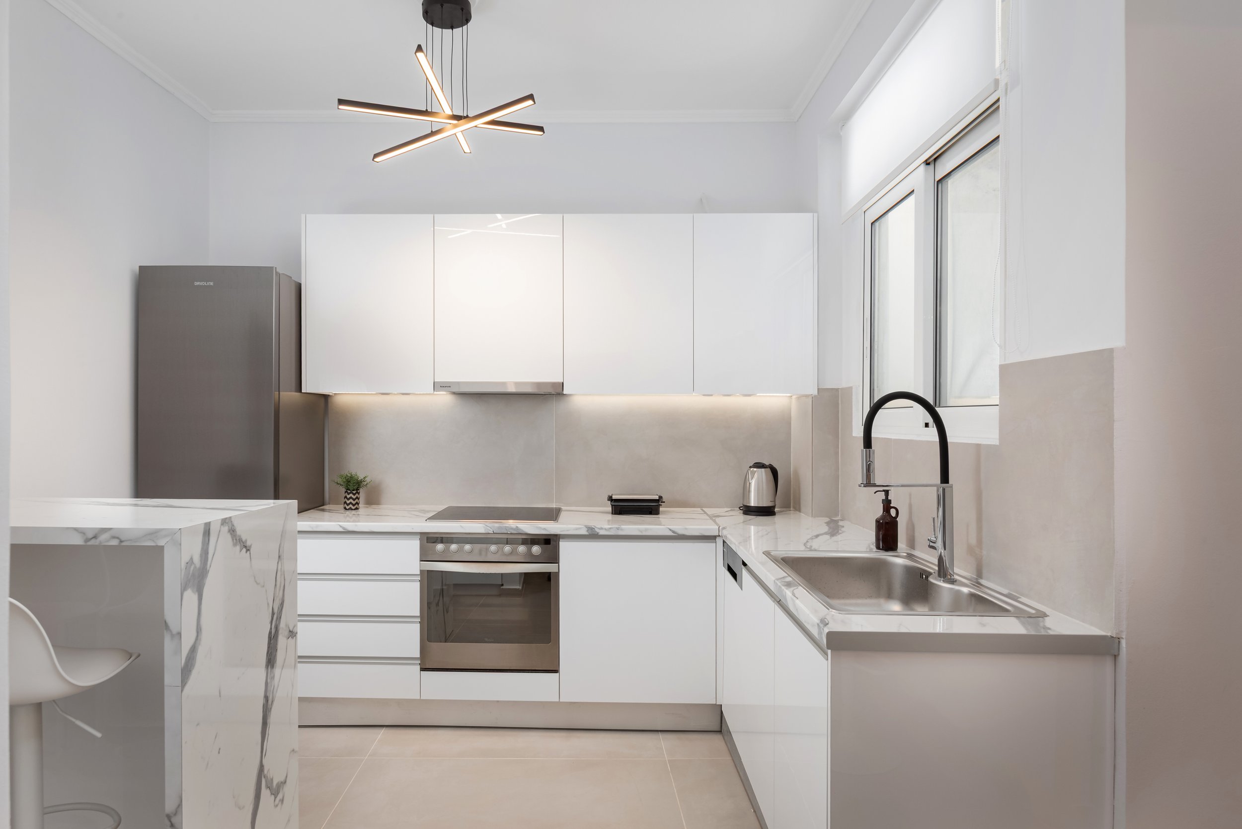 Modern minimalist kitchen with white cabinets, marble countertops, stainless steel appliances, a small potted plant, and a large window letting in natural light.