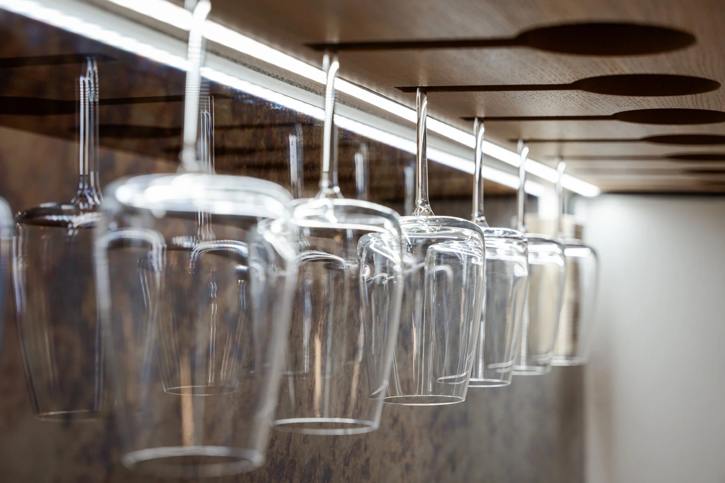 A modern bar area with two wooden shelves, hanging wine glasses, and white bar stools.