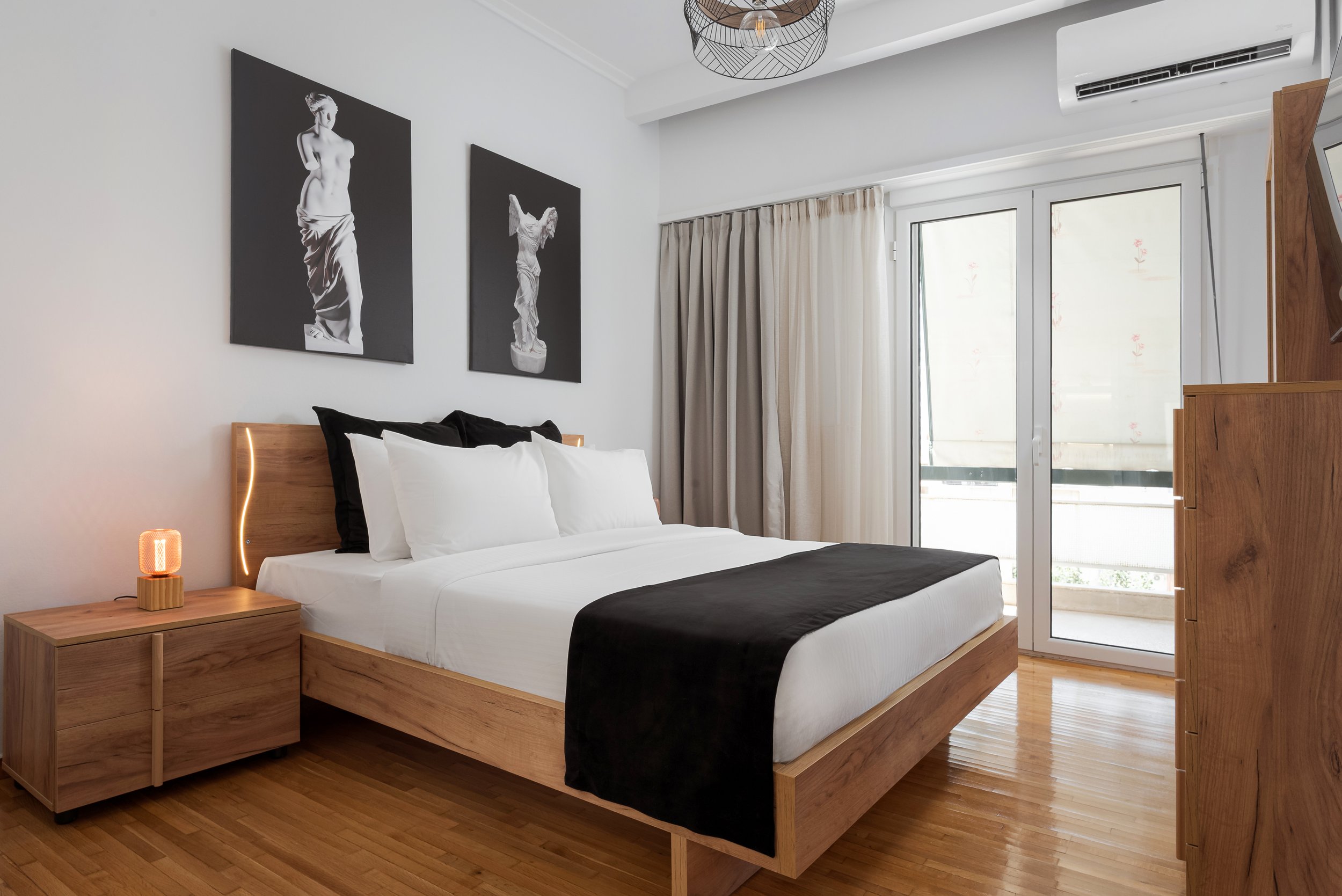 Modern bedroom with a wooden bed, white linens, black and white artwork on the wall, a wooden nightstand with a small lamp, beige curtains, and glass doors leading to a balcony.