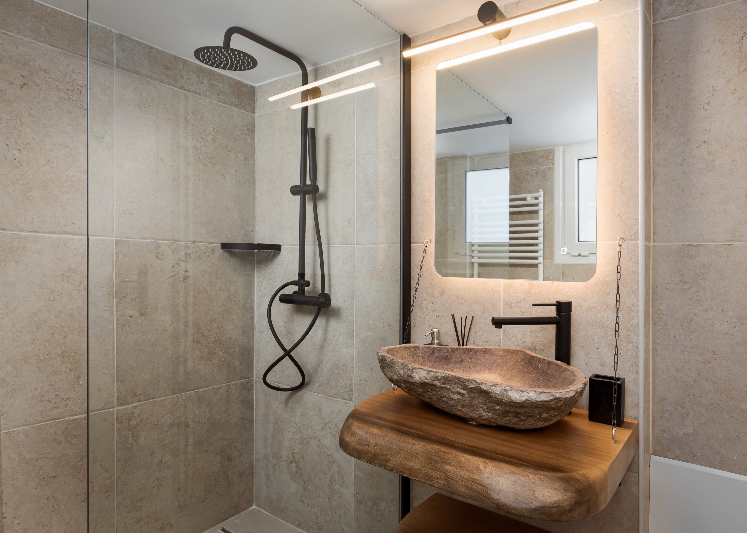 Modern bathroom with a walk-in shower featuring a black showerhead and fixtures, and a stone vessel sink on a wooden countertop with a large backlit mirror above, beige tiled walls, and a small window.