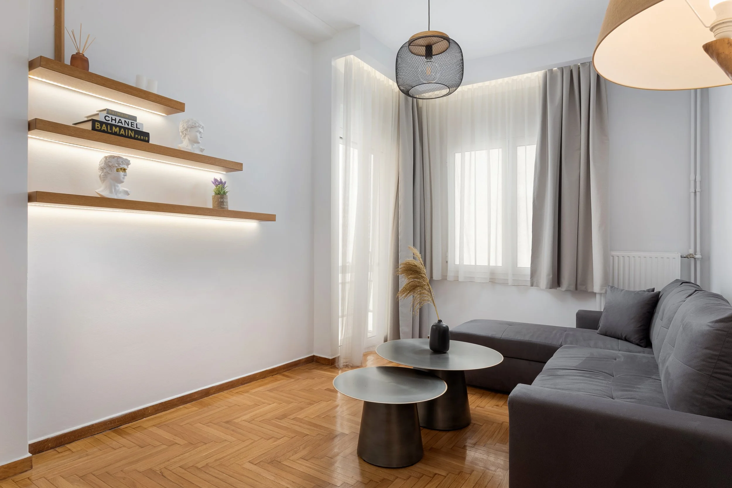 Modern living room with a gray couch, wooden floor, and floating shelves with decorative busts and books, illuminated by natural light through sheer curtains.