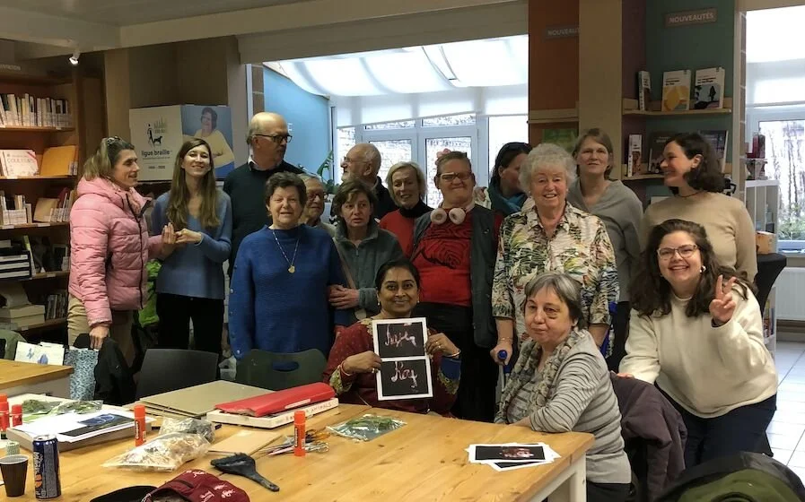 Group of diverse people gathered in a library or bookstore, some sitting at a table with books and craft supplies, others standing behind, smiling and posing for a photo.