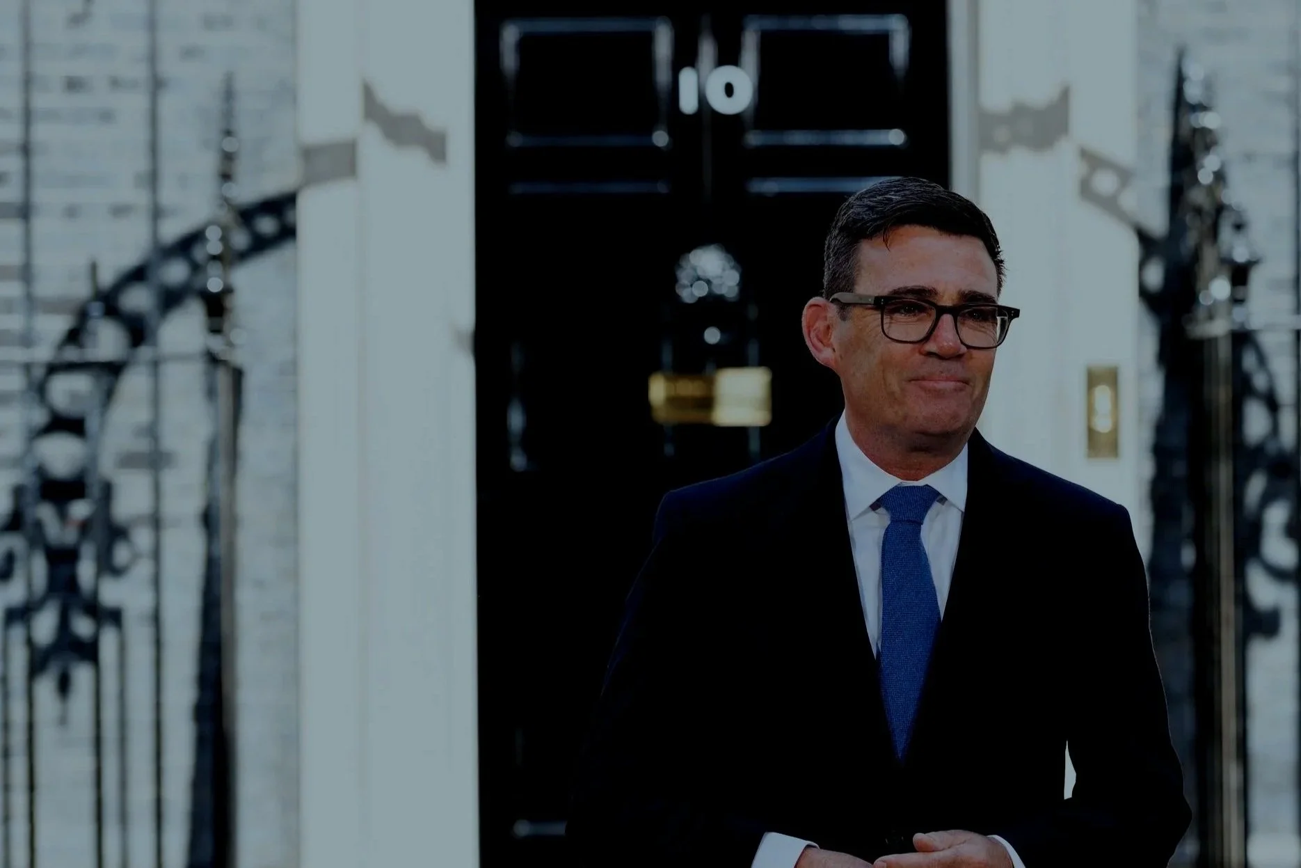 A man wearing glasses, a dark suit, white shirt, and blue tie, standing outside a building with white brick walls and black gates, in front of a black door with a gold mail slot.