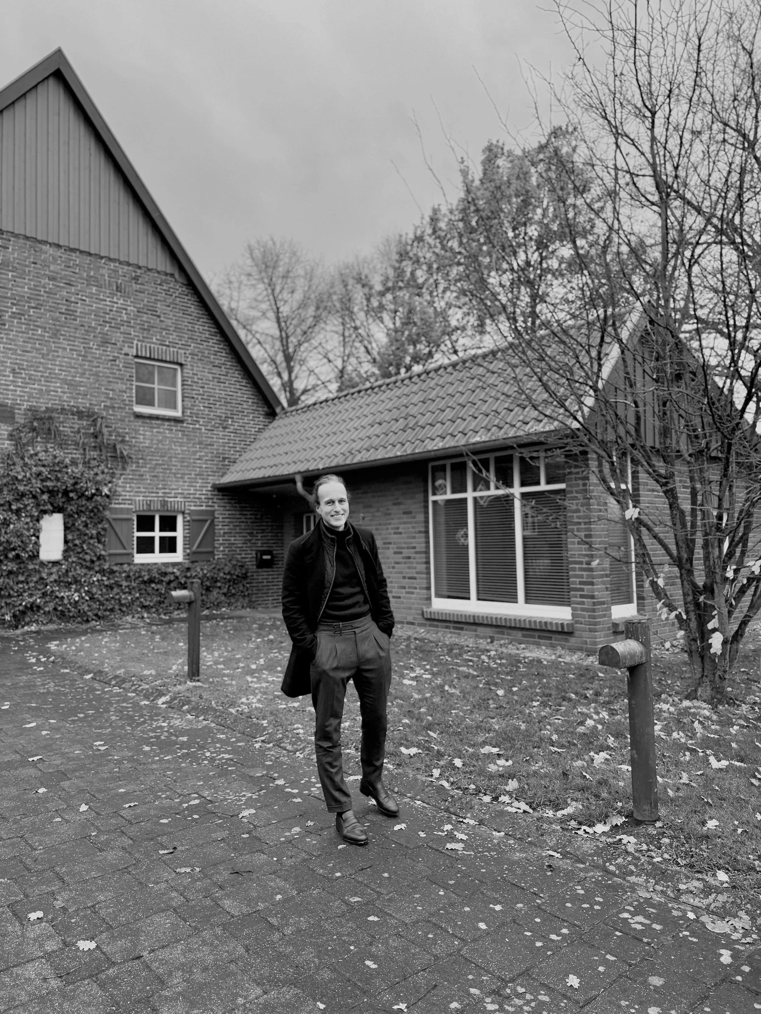 Ein Mann in dunkler Jacke, schwarzem Pullover, schwarzen Hosen und Stiefeln steht auf einer gepflasterten Straße vor einem Haus mit roten Ziegeln und großen Fenstern. Das Haus hat ein Satteldach, im Hintergrund sind kahle Bäume sichtbar, es ist bewölkt.