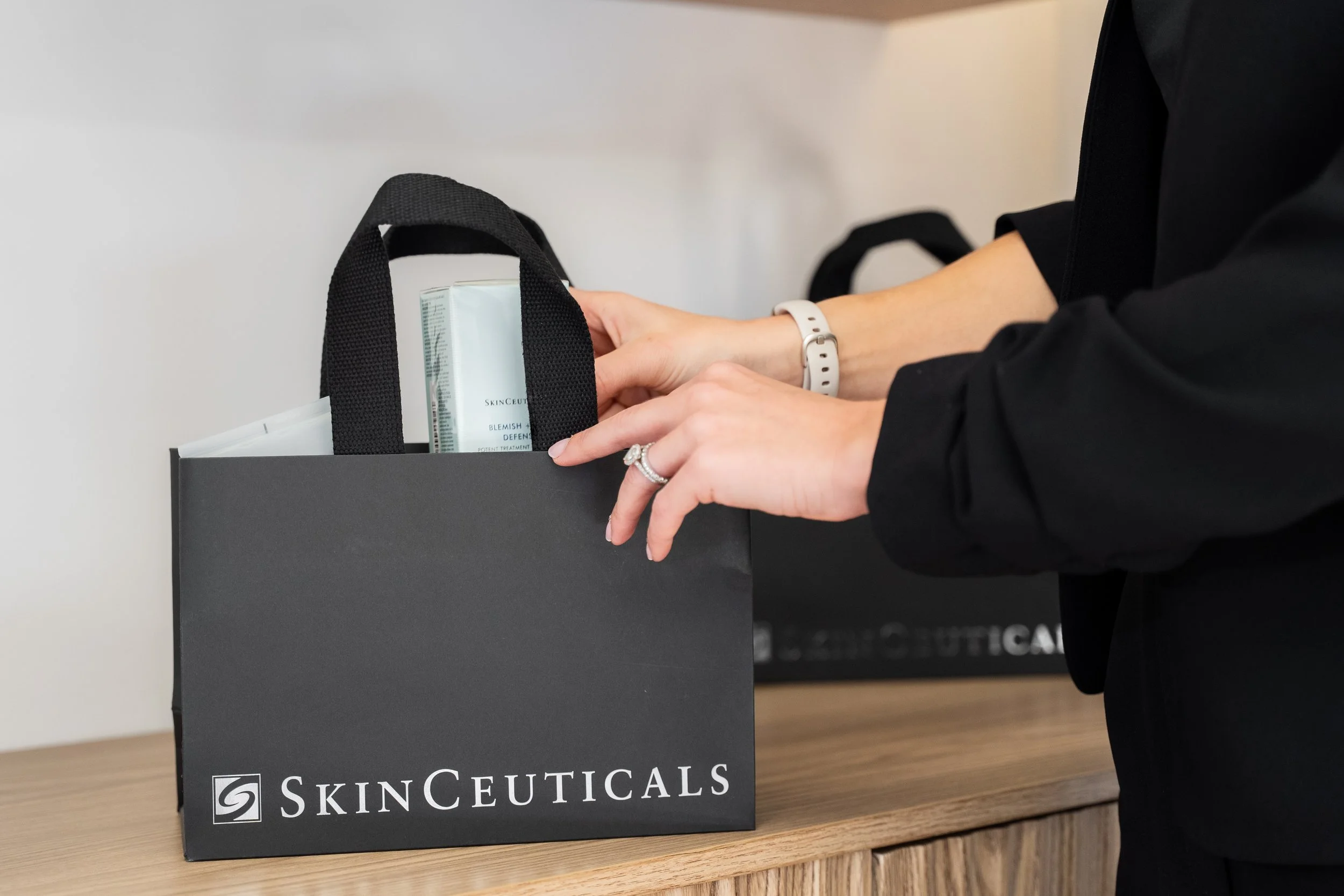 Person placing skincare products into a black SkinCeuticals shopping bag on a wooden surface.