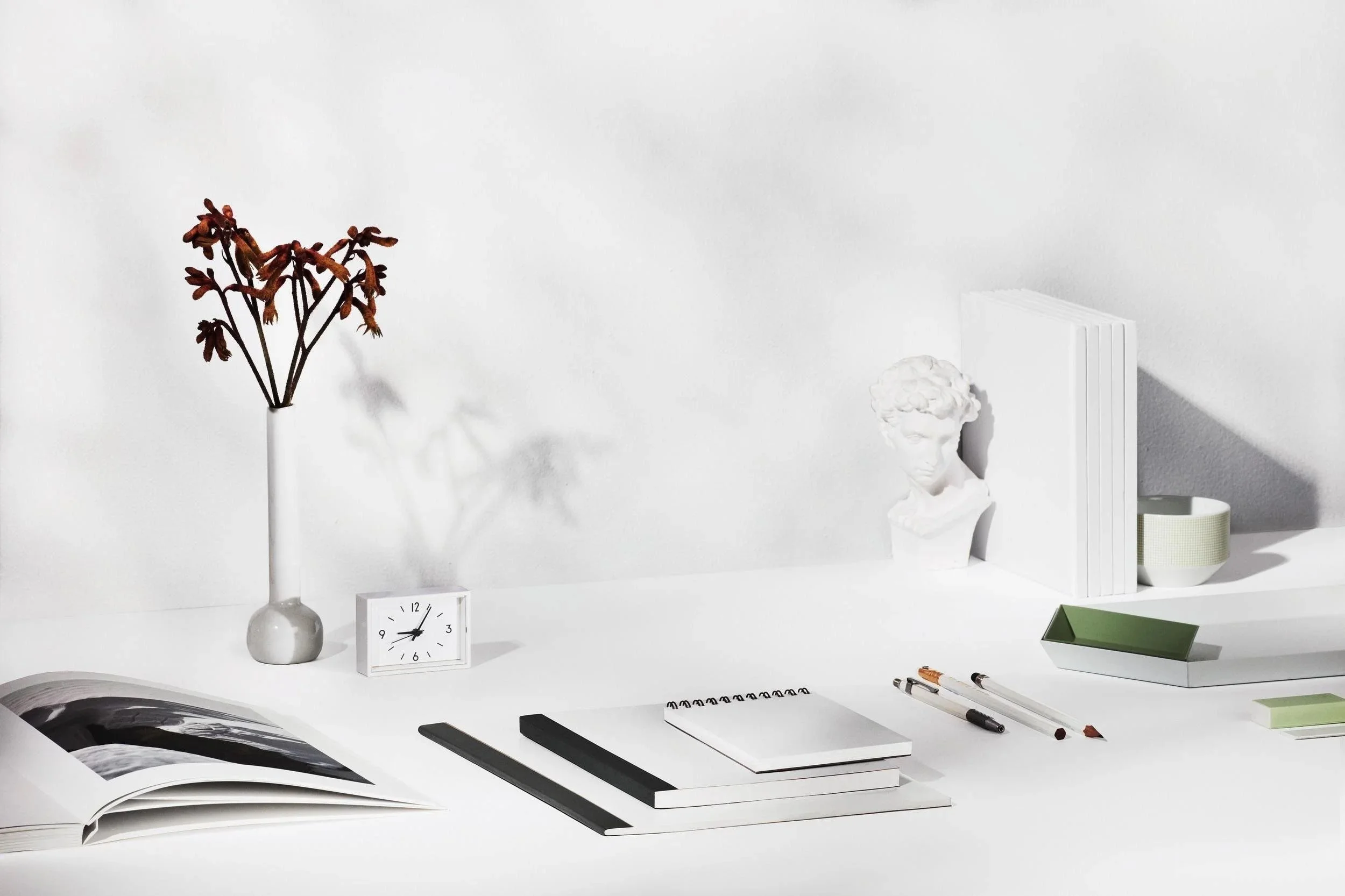 Minimalist white desk with a white vase holding dried flowers, a small white clock, an open book, notebooks, pens, a green tray, and decorative objects including a white bust and a small white book with green accents.